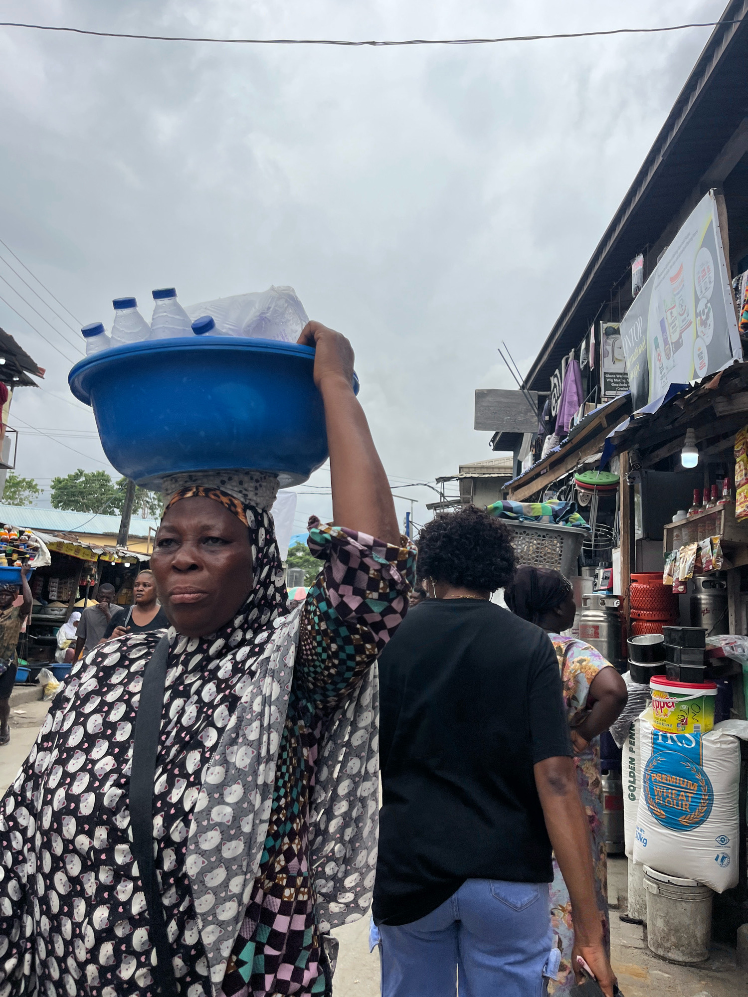 In a bustling marketplace in Ajah, Nigeria, a woman carries a blue basin filled with bottled water and other items on her head. She wears a patterned headscarf and a black and white patterned tunic with a black crossbody bag. Behind her, a person with curly hair wears a black t-shirt and light blue jeans, while another individual in a floral dress stands nearby. The market is lined with stalls selling various goods, including bags of flour, containers, and other merchandise. The scene is captured under a cloudy sky, suggesting an overcast day. The overall atmosphere is one of everyday commerce and activity.