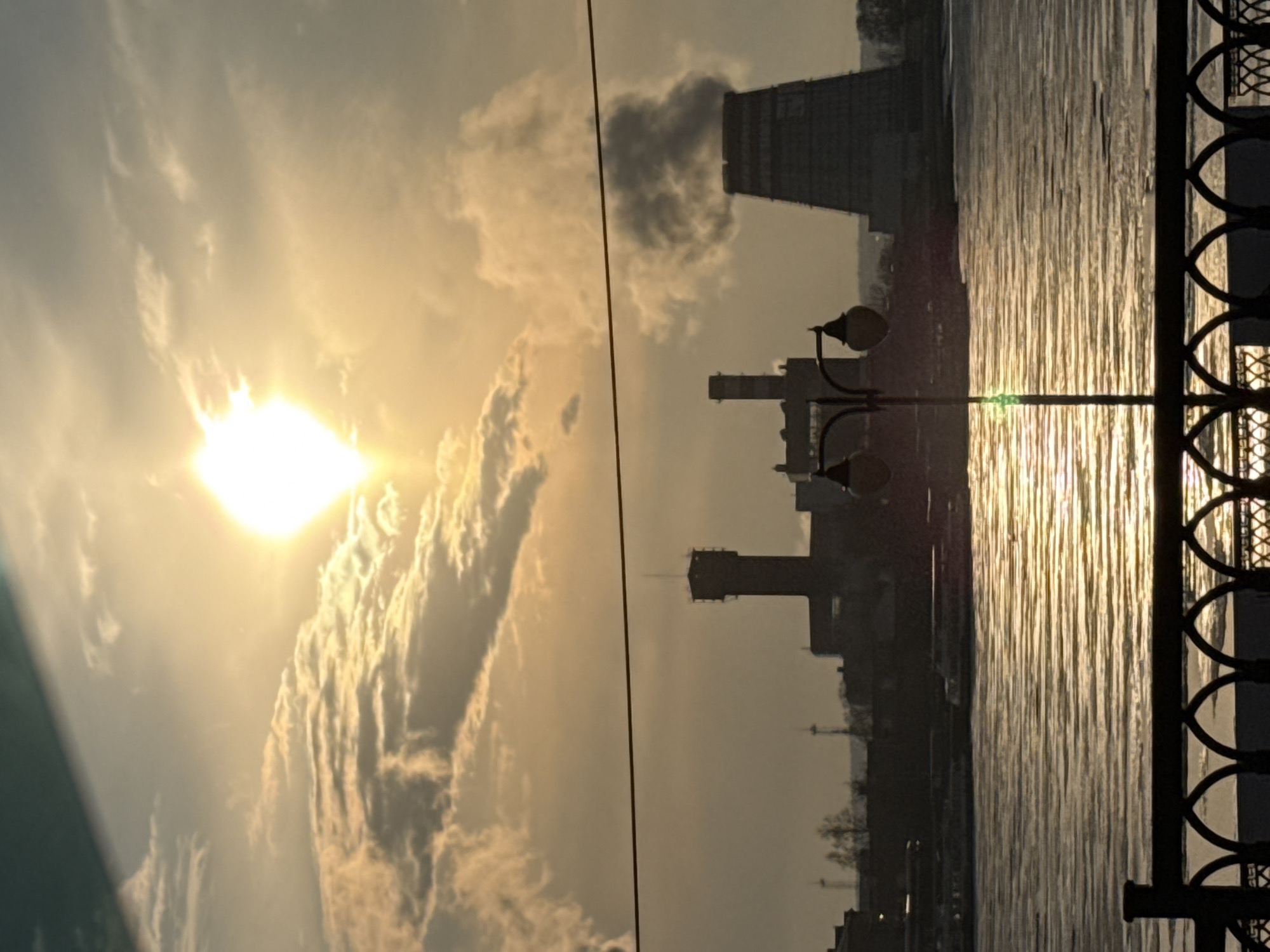 The image depicts a sunset scene over the Izhevsk Pond in Izhevsk, Russia. The sun, partially obscured by clouds, casts a bright glow over the sky and reflects on the water's surface. In the background, the silhouettes of industrial buildings and towers are visible. A decorative metal railing runs along the right edge of the frame, and a streetlight is positioned closer to the center of the image. Smoke can be seen coming from one of the buildings, which appear to be the Izhevsk Thermal Power Plant. The overall scene is urban and industrial, but also serene due to the sunset.