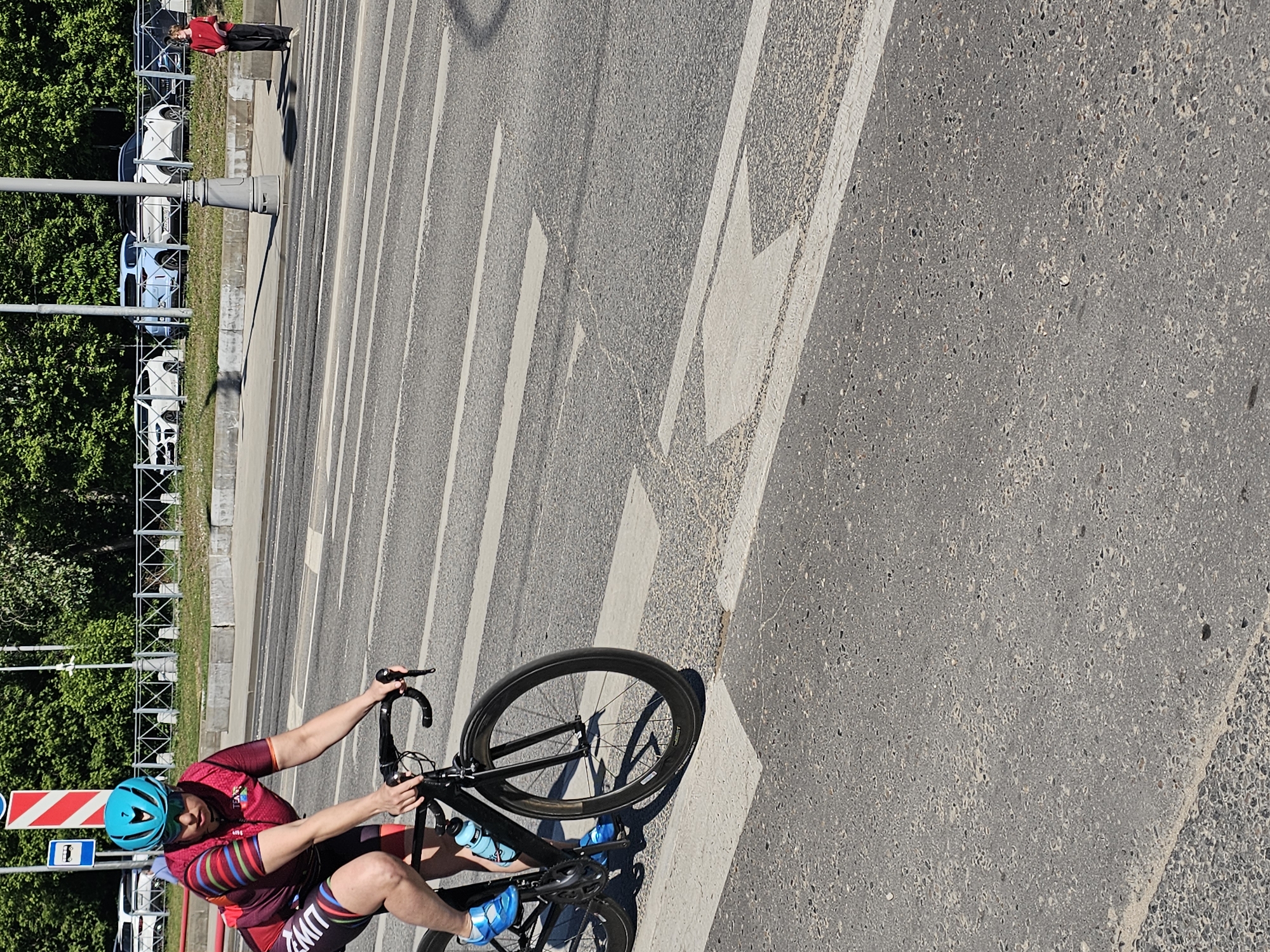This image captures an urban street scene in Moscow, Russia, on a bright, sunny day. The primary subject is a cyclist, presumably male, positioned in the foreground on a black road bicycle. He is wearing a distinctive red, black, and striped cycling jersey and shorts, a teal helmet, and bright blue cycling shoes. Two blue water bottles are visible on his bicycle frame. The cyclist appears to be either stopped or in the process of starting/stopping, with his hands on the handlebars and looking forward to the right.
In the background, on the left side of the road, another person wearing a red shirt and dark pants stands on a sidewalk. Behind them, several cars (including white and light blue vehicles) are parked along a grassy area, separated from the sidewalk by a metal barrier. Lush green trees line the very far background.
The road itself is wide, made of asphalt, and features clear white painted markings, including a zebra crossing and a large directional arrow. Strong shadows cast by the bicycle, a street lamp, and the person in the background indicate bright daylight and clear weather. A red and white striped road sign, along with a blue sign depicting a pedestrian or child, are visible near the cyclist. Text partially visible on the cyclist's shorts reads "LIVWZ".