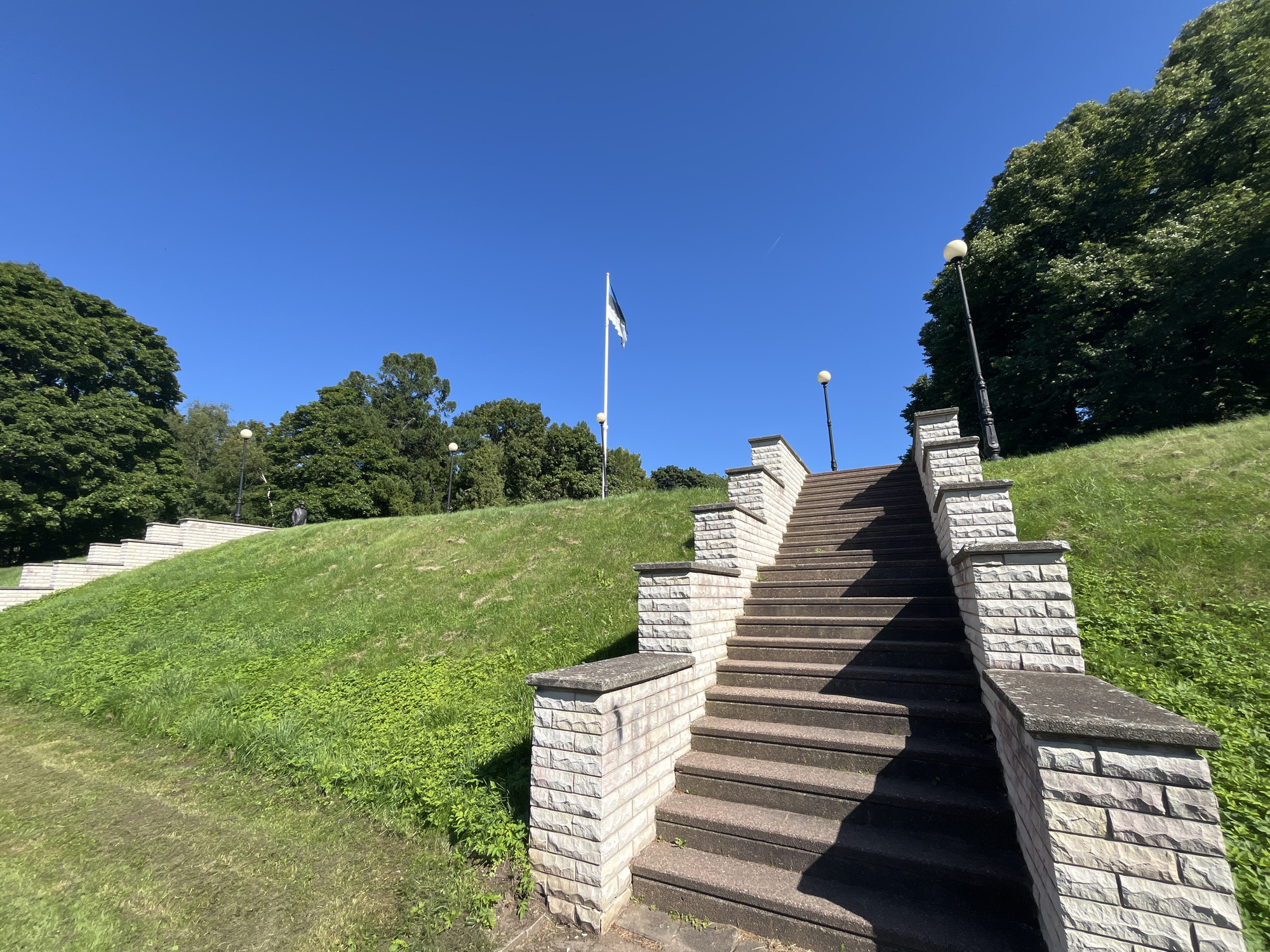 The image depicts a wide stone staircase leading up a grassy hill under a clear blue sky. The staircase is bordered by low stone walls topped with flat grey caps, constructed from light-colored, stacked rectangular stones. The steps themselves appear to be made of a darker, possibly concrete or stone material, and are worn with use. Green grass covers the slopes of the hill, with patches of smaller green plants growing along the base of the stairs. Several lampposts with spherical lights are visible along the staircase and on the higher ground. In the distance, a flagpole flies the Estonian flag, which consists of three horizontal stripes of blue, black, and white. Lush green trees frame the scene on the left and right, with a denser concentration of trees on the far right, creating a shaded area on the hill. The overall impression is a well-maintained public space, likely a park or memorial, in Toila, Estonia.