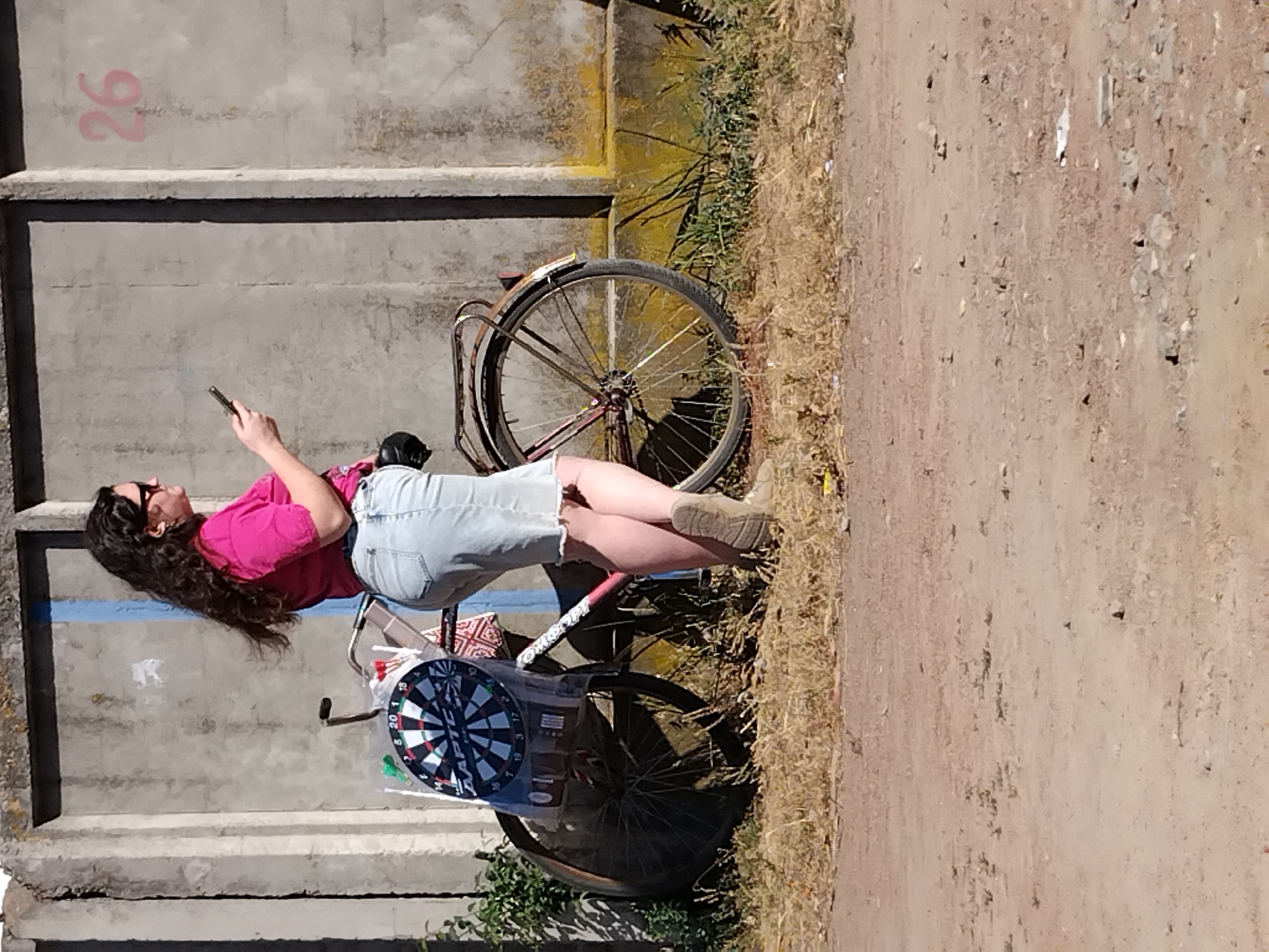 In Irshansk, Ukraine, a woman with long, dark, wavy hair and sunglasses stands beside a bicycle. She is wearing a pink short-sleeved shirt and a light blue denim skirt. The bicycle has a dartboard attached to its frame and a small red taillight. The woman is holding a mobile phone in her right hand, and her left hand is resting on the bicycle's frame. The scene takes place outdoors, with a concrete wall behind the woman and the bicycle, and dry grass and dirt in the foreground. The wall has the number "26" spray-painted in pink on its surface.