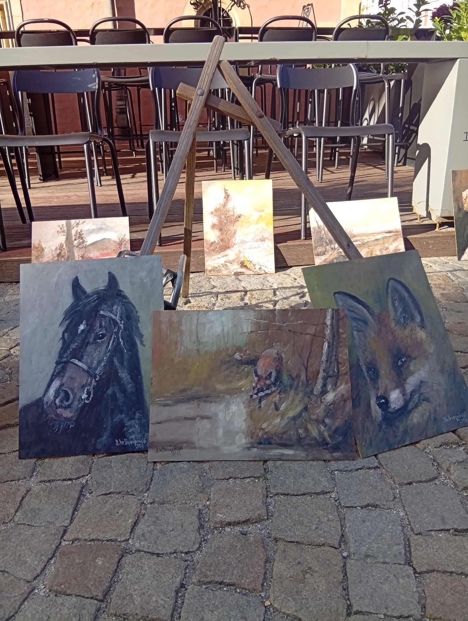 The image depicts an outdoor display of paintings on a cobblestone ground, likely in Wrocław, Poland, given the style of the paintings and the context of street art displays often seen in European cities. In the foreground, several paintings are arranged. One is a portrait of a black horse, facing left, with a detailed bridle. To its right is a painting of a fox's head, also facing left, with intense eyes. Between these two, and slightly behind, is a painting that appears to show a bird in a wooded environment. In the background, a wooden easel supports at least two more paintings, one showing a landscape with trees and a hazy sky, and another with a similar rustic theme. Behind the easel and paintings, rows of dark chairs and tables are visible, suggesting an outdoor seating area, possibly of a cafe or restaurant, with a pinkish-toned building facade in the far background. The lighting indicates it is daytime, with sunlight casting shadows and highlighting the textures of the cobblestones and the painted surfaces.