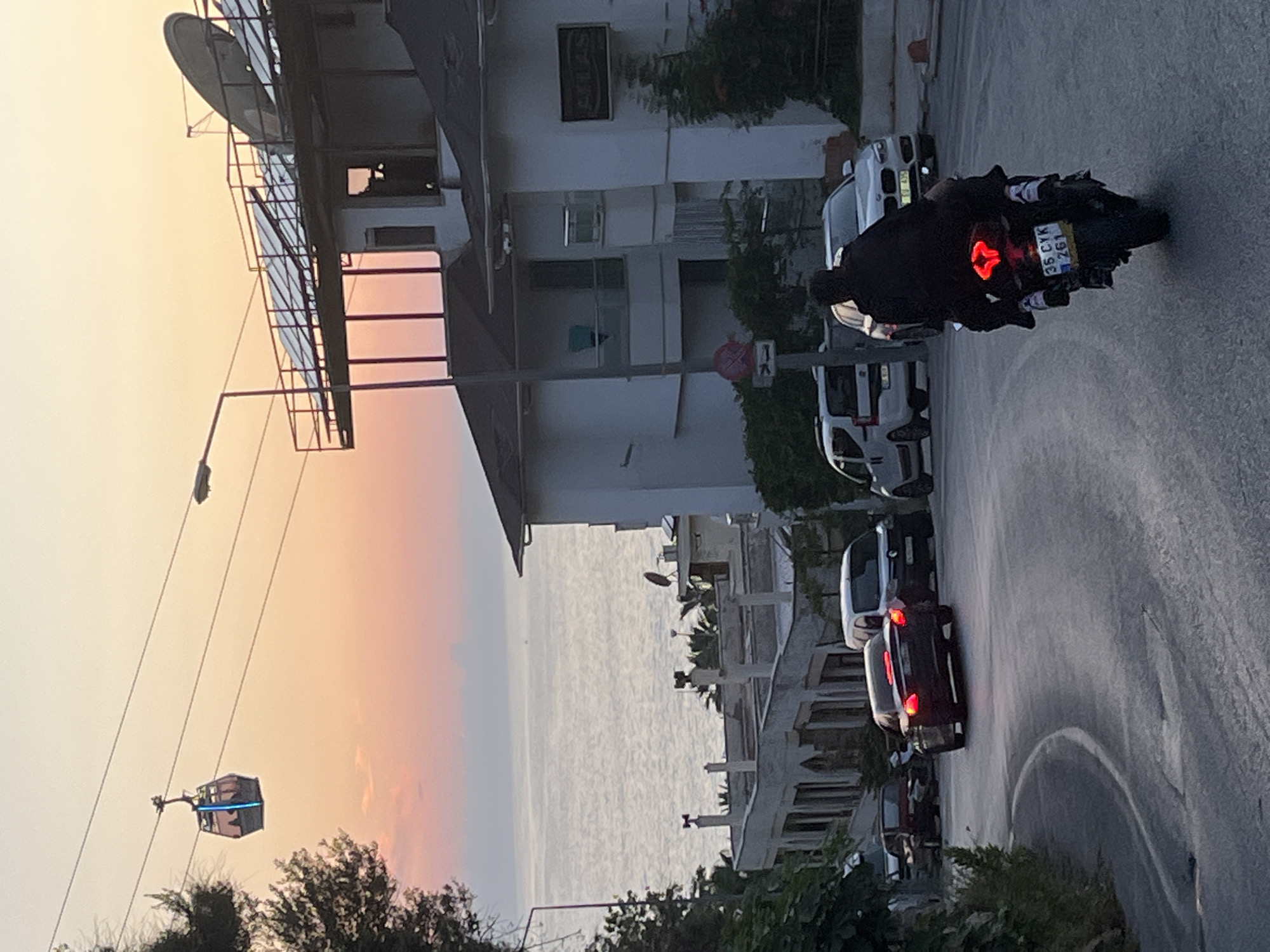 The image shows an urban scene with a street winding past buildings. On the right side, a motorcyclist with a passenger are visible on a motorcycle with the license plate "35 CYK 261". Several parked cars line the street. In the background, the sea stretches out to the horizon, reflecting the colors of the sky. A cable car is visible to the left, suggesting the location is Alanya, Turkey. Buildings of various heights are present along the street, including what looks like residential buildings. The sky transitions from a light orange near the horizon to a lighter color overhead, indicating either sunrise or sunset. Vegetation is visible in the foreground. The road surface is dark and shows signs of wear.