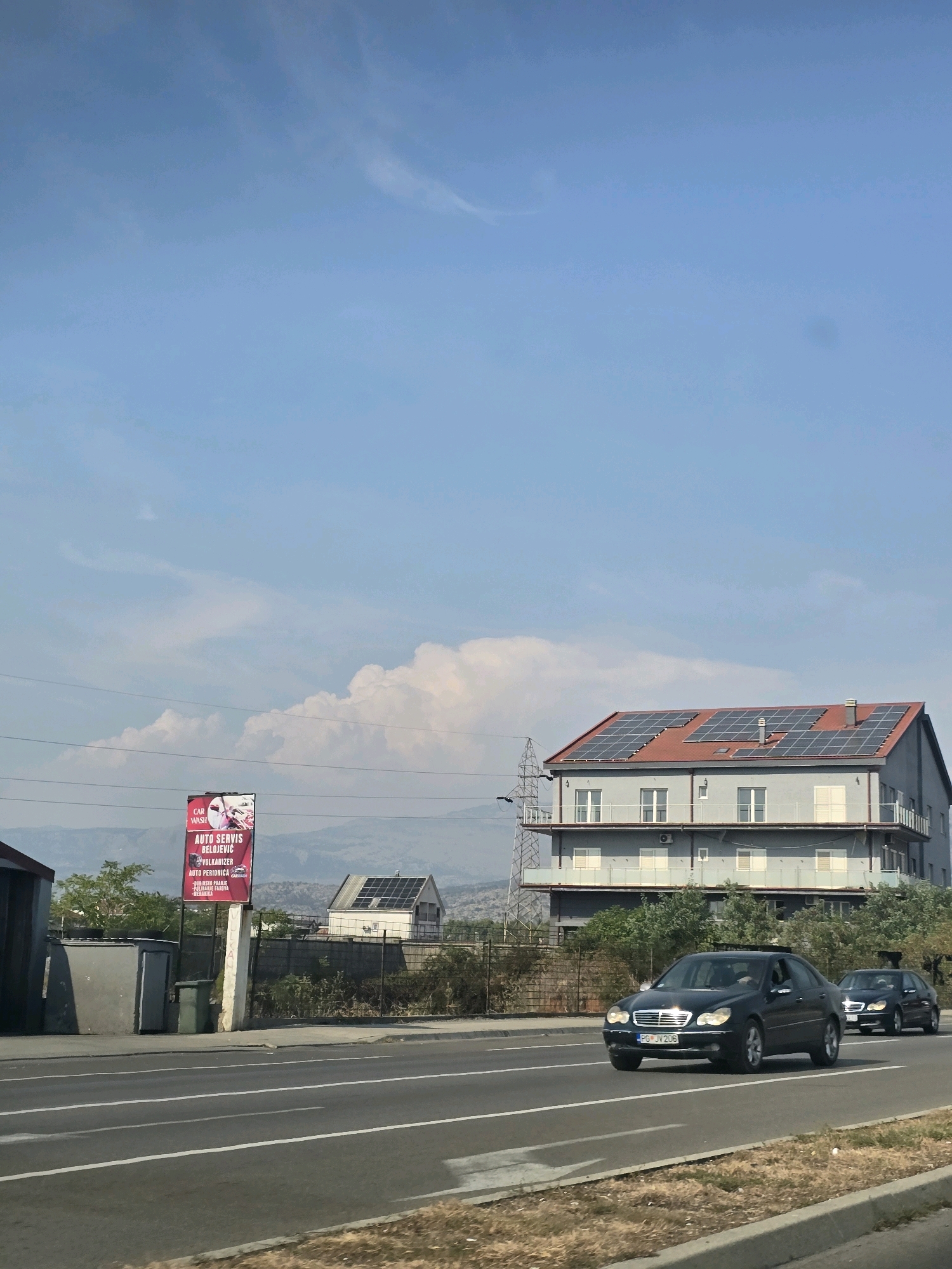 This image shows a street scene in Podgorica, Montenegro. In the foreground, two dark-colored sedans are driving on a multi-lane asphalt road. The car in the left lane has the license plate "PG JV 206". In the middle ground, there is a row of buildings and trees. One building, a large, modern structure with a red roof covered in solar panels, is prominent. To the left of this building, a billboard advertising "AUTO SERVIS BELOJEVIĆ" is visible, along with other services such as "CAR WASH", "VULKANIZER", "AUTO PERIONICA", and "POLIRANJE FAROVA - GENERIKA". Further in the background, mountains are visible under a clear blue sky with scattered clouds. The environment appears to be a suburban or industrial area on the outskirts of the city, indicated by the mix of commercial buildings, road infrastructure, and natural landscape. No people are clearly visible in the image.