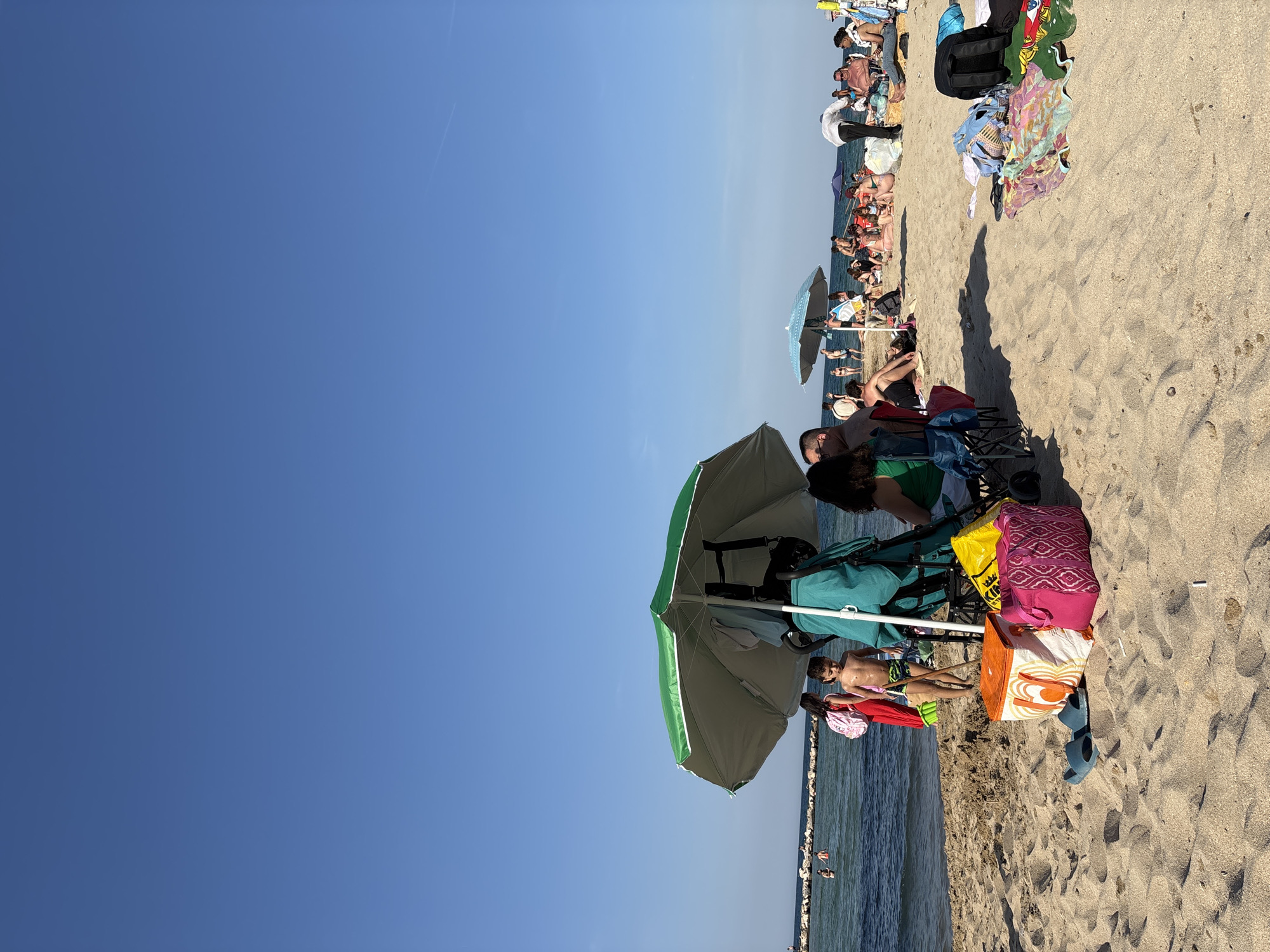 The image shows a crowded beach on a sunny day. Several people are sitting in chairs under umbrellas. Other people are sitting on blankets. The beach is sandy. The water is blue.