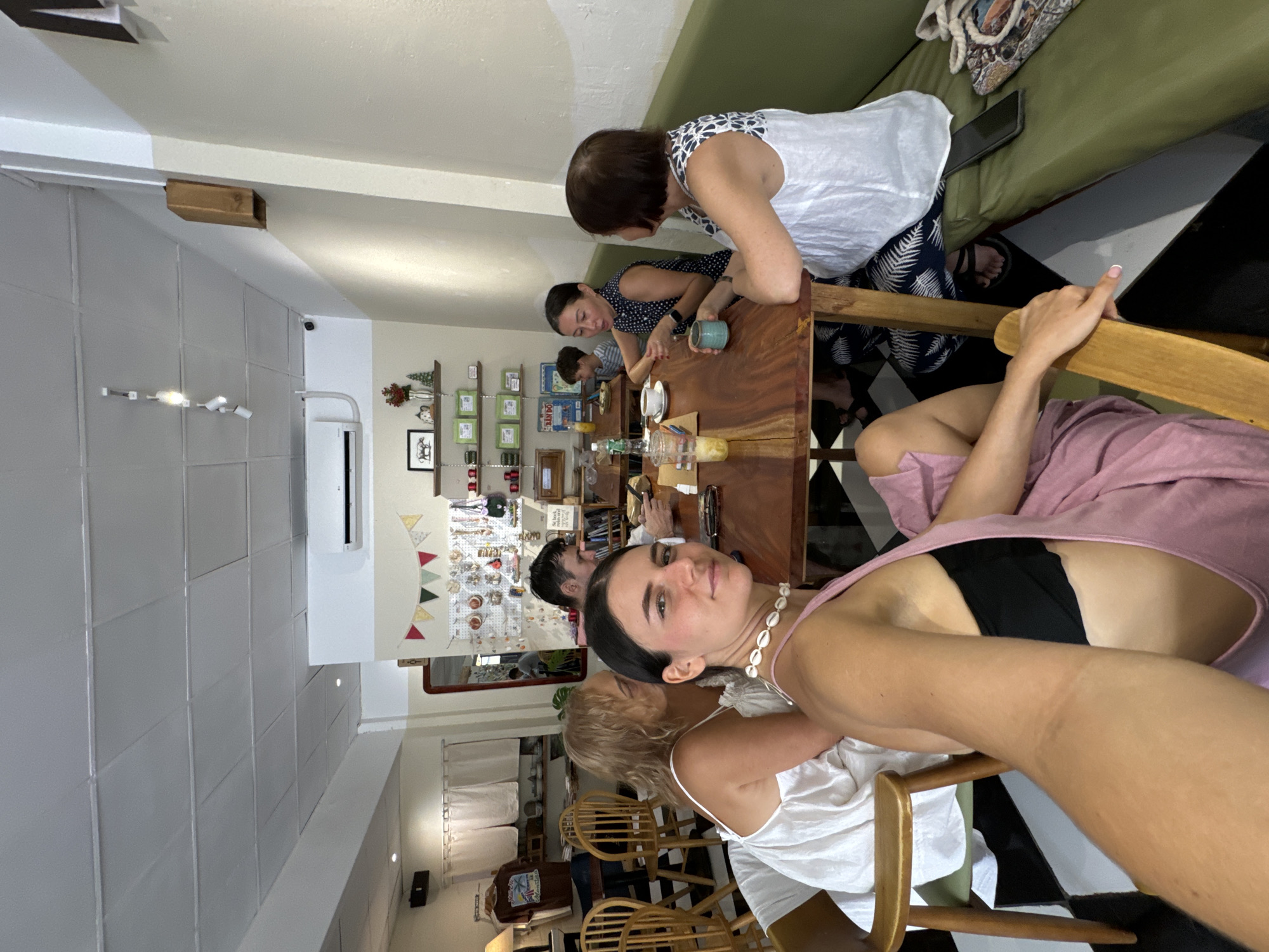 The image captures an indoor scene, likely a cafe or restaurant, filled with natural light and a casual atmosphere. The setting features light-colored walls, a ceiling with rectangular panels, and a floor partially covered in a black and white checkerboard pattern. A long, dark wooden table serves as the central gathering point, flanked by a long green upholstered bench along one wall and individual wooden chairs.
Several individuals are present, engaging in various activities. In the foreground, a woman with dark hair, wearing a pink top and a shell necklace, looks directly at the camera, seemingly taking a selfie or being photographed. Her black bra is visible beneath her top. Beside her, a blonde-haired woman in a white top is partially visible, looking away from the camera. Around the wooden table, a woman with short dark hair in a patterned white and black top is seated, looking down, while another woman with dark hair in a polka-dotted top holds a teal cup, also focused downwards. A young boy is partially visible between them. Further down the table, another person is engrossed in a mobile phone. A total of about six to eight people can be observed within the frame.
The table is adorned with various drinks in cups and glasses. Decorative elements include small shelves on the back wall holding ornaments and framed pictures, and a string of colorful triangular pennants. An air conditioning unit and a small CCTV camera are mounted on the wall. A rectangular mirror reflects parts of the room. The overall impression is of a daytime gathering in a comfortable, decorated cafe. No legible text is clearly visible in the image. The location context indicates this scene is in Phu Thuy Ward, Vietnam.