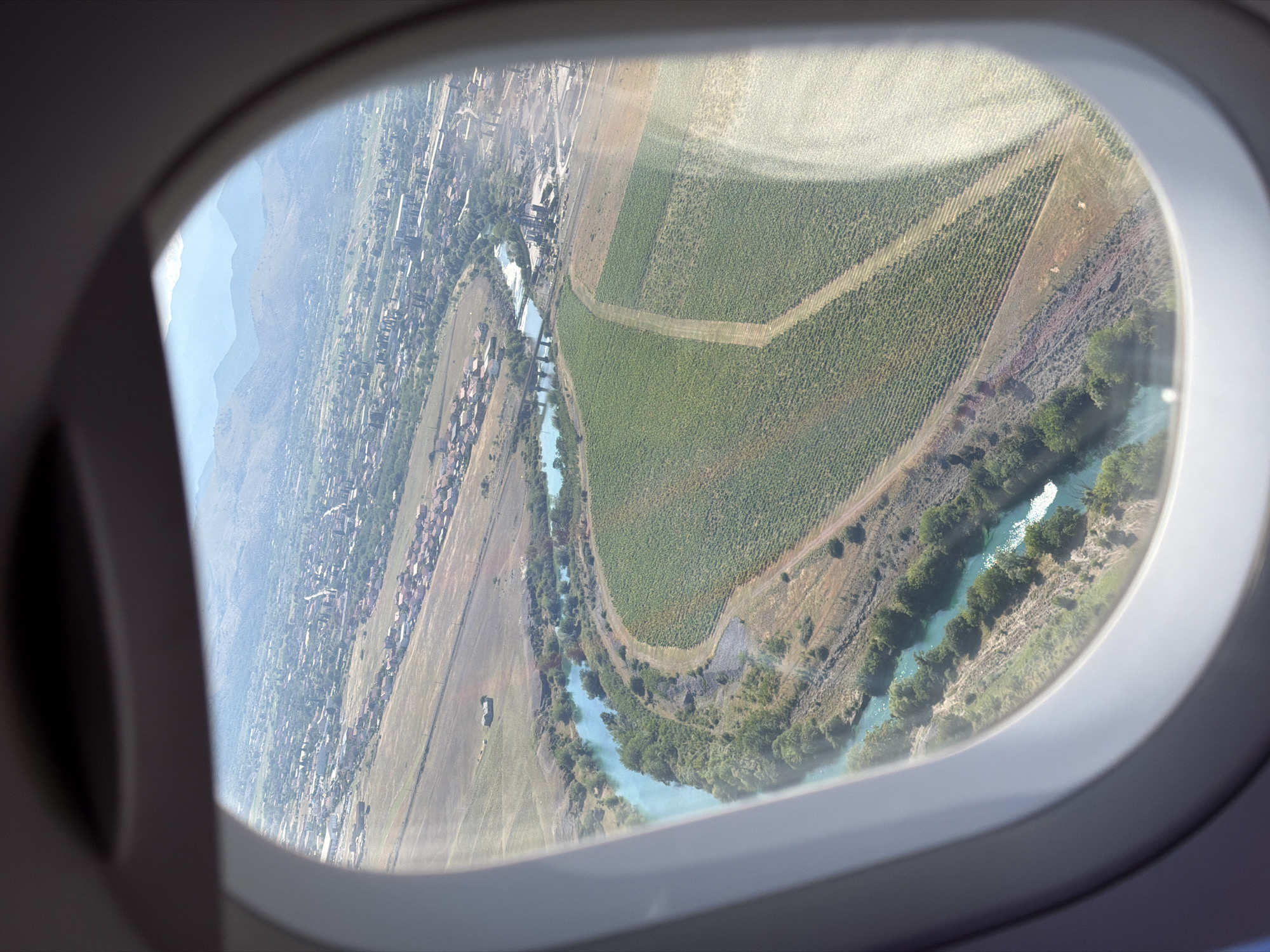 The image is a high-angle aerial view taken through an airplane window, overlooking the city of Donja Cijevna, Montenegro. The landscape features a winding, turquoise river with lush green vegetation along its banks. Adjacent to the river are fields of crops, appearing to be vineyards, with distinct rows of plants. Further inland, a settlement with red-tiled roofs is visible, interspersed with trees. In the background, rolling hills and mountains stretch towards the horizon under a hazy, light-blue sky. The scene is dominated by natural elements such as the river, vegetation, and mountains, alongside man-made structures including the village and agricultural fields. There are no visible people or specific actions depicted.