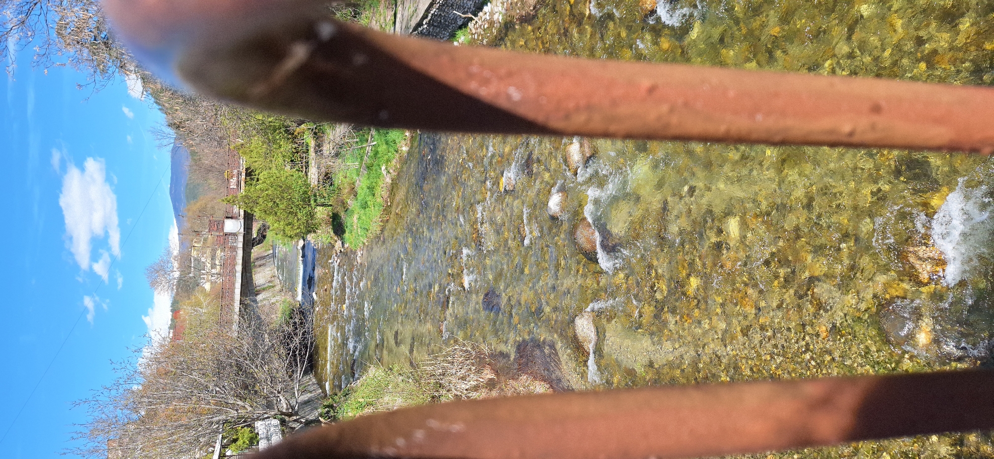 The image shows a view of a river in Blagoevgrad, Bulgaria, looking downstream from what appears to be a bridge with rusty railings. The river is shallow and clear, with visible rocks and pebbles on the riverbed. The water is flowing rapidly, creating small rapids and splashes. On the left bank of the river, there is vegetation including grass and trees. In the background, buildings and mountains are visible. The sky is blue with scattered clouds. The rusty bridge railings are prominently featured in the foreground, obscuring the view somewhat.