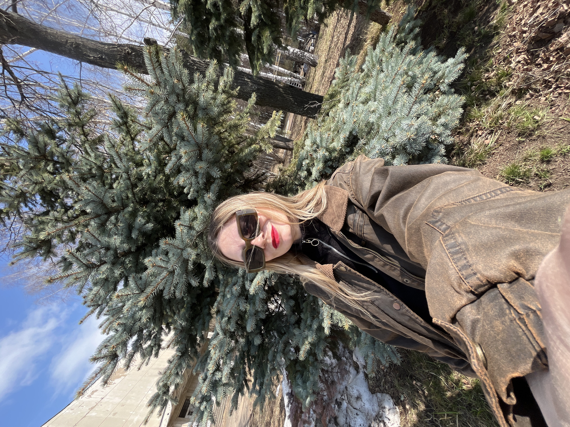 The image is a bright outdoor selfie featuring a young woman in Chaikovsky, Russia, taken on a sunny day during late winter or early spring. The woman, with long blonde hair, is in the foreground, wearing dark, rectangular sunglasses and striking red lipstick. She is dressed in a rugged, dark brown jacket with a corduroy collar, possibly made of waxed cotton or distressed leather, over a black top with a distinctive silver zipper pull.
The background is dominated by lush blue-green coniferous trees, likely spruces, indicating a park or natural setting. Beyond these evergreens, the bare branches of deciduous trees are visible against a clear, bright blue sky with a few wispy white clouds. The ground shows patches of dry grass and remnants of melting snow, confirming the transitional season. A light-colored building can be partially seen in the far left background, adding an urban or suburban element to the natural landscape. The bright sunlight casts clear shadows, emphasizing the textures of the trees and the woman's attire. The activity is clearly a self-portrait, with the woman looking directly towards the camera. There is no visible text in the image.