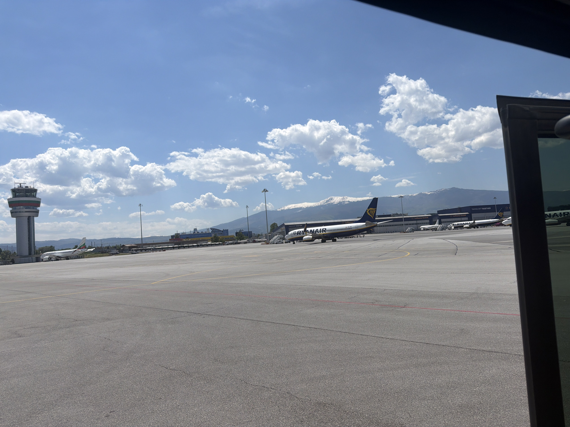 The image depicts an airport scene, likely at Sofia Airport in Bulgaria, given the control tower and the presence of a Ryanair aircraft. The sky is blue with scattered cumulus clouds, and mountains are visible in the distance. The foreground consists of the airport tarmac, with painted yellow and red lines. A Ryanair aircraft is positioned on the tarmac, and several other airplanes are visible in the background. In the far left, there's an airport control tower. An open window or frame partially obscures the right side of the image. No people are clearly visible.