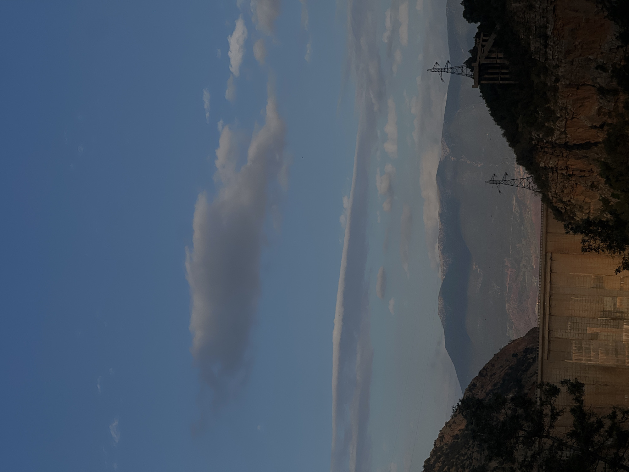 The image shows a partial view of the Bin El Ouidane Dam in Morocco, nestled within a mountainous landscape. The concrete dam wall is visible on the right, adjacent to a rocky slope covered in vegetation. Two electrical transmission towers are positioned on the cliff's edge, above a mountain range in the background. The sky, adorned with scattered clouds, fills the left side of the frame.