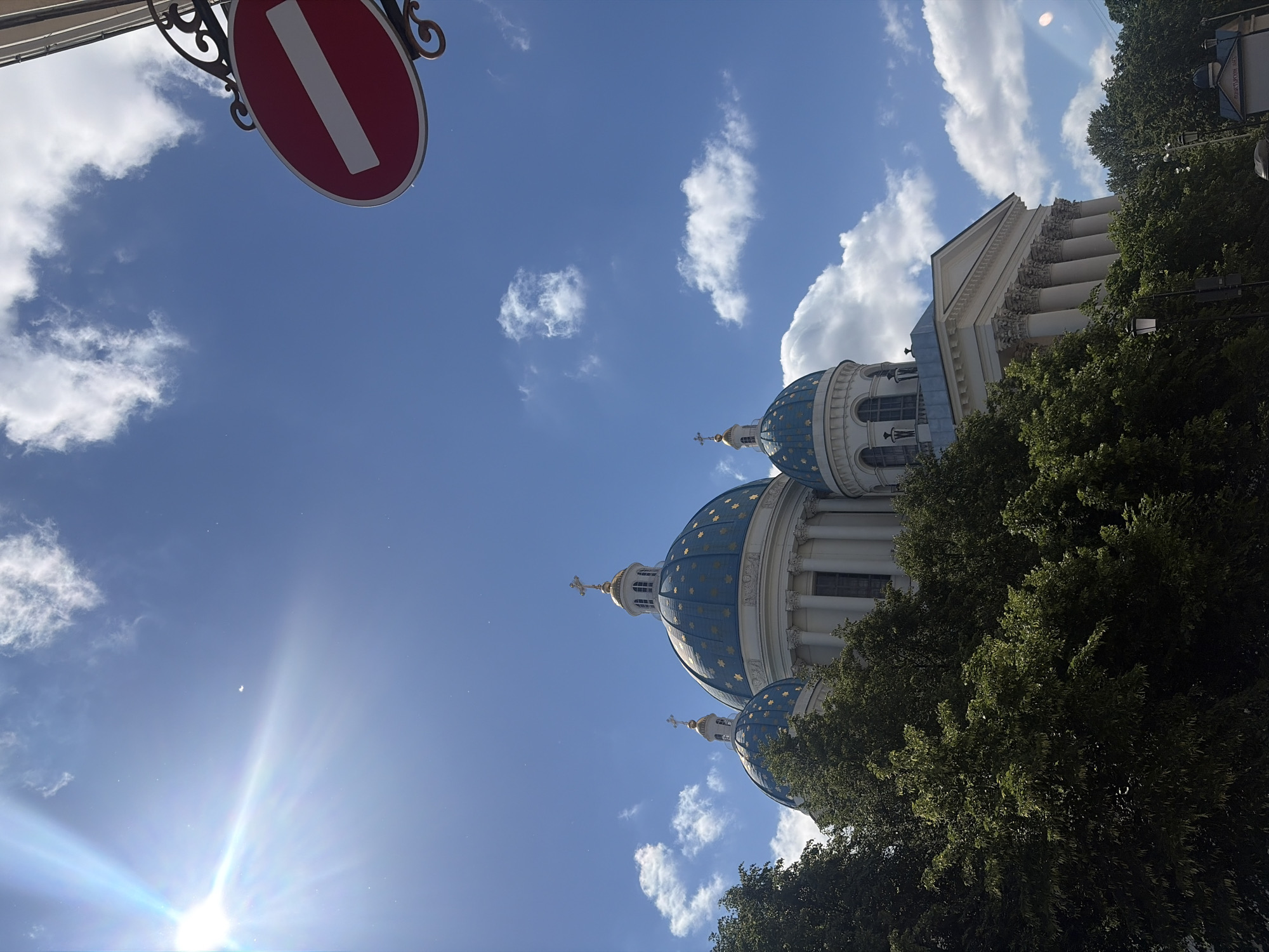 This is a low-angle, outdoor, daytime photograph of the Trinity Cathedral in Saint Petersburg, Russia. The cathedral's distinctive blue domes, adorned with gold stars, are visible against a bright blue sky with scattered white clouds. Sunlight streams in from the upper left corner, creating a lens flare effect. In the foreground, lush green trees partially obscure the lower levels of the cathedral.
A red and white "no entry" sign hangs from an ornate black metal bracket in the upper right quadrant of the image. The architecture of the cathedral is classical, featuring columns and a pediment on its main facade. There are no people visible in the frame. The environment appears to be urban, with streetlights and a small blue-domed structure with text visible in the lower right corner, suggesting a civic or religious complex. The overall impression is one of grandeur and historical significance, set against a clear, sunny day.
