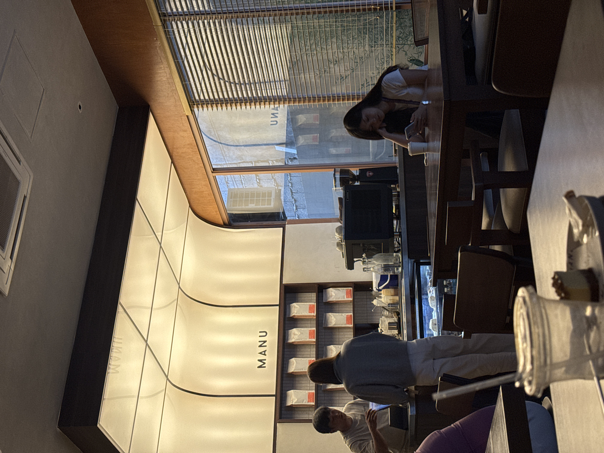 A high-angle interior shot shows a cafe in Davao City, Philippines. The cafe has a bright, modern design with the name "MANU" displayed on a lighted wall. Customers are seated at a long wooden table; one woman is using her phone, and two employees stand behind the counter. Merchandise is shelved behind the counter, and daylight streams in through the window blinds.