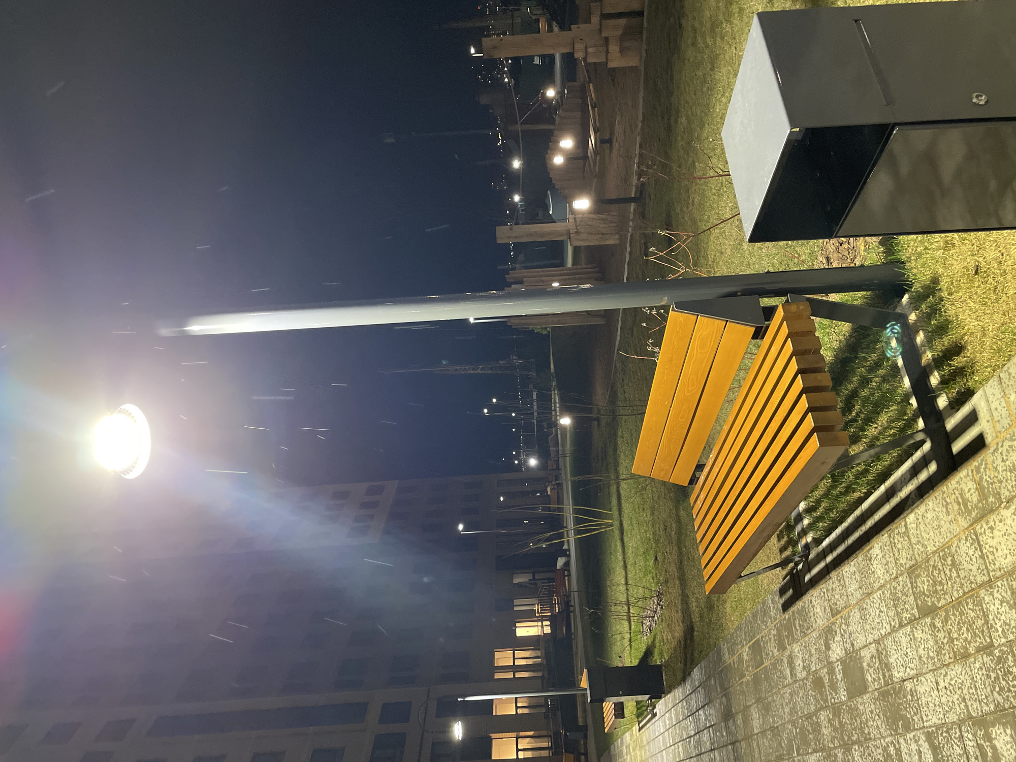 The image depicts a night scene in Izhevsk, Russia. It is snowing lightly. A yellow wooden bench sits on a green lawn next to a gray brick walkway. A modern black trash receptacle is situated near the bench. A tall streetlight illuminates the area, casting a strong glare. In the background, a multi-story building with illuminated windows is visible, along with other streetlights and what appears to be a decorative wooden structure.