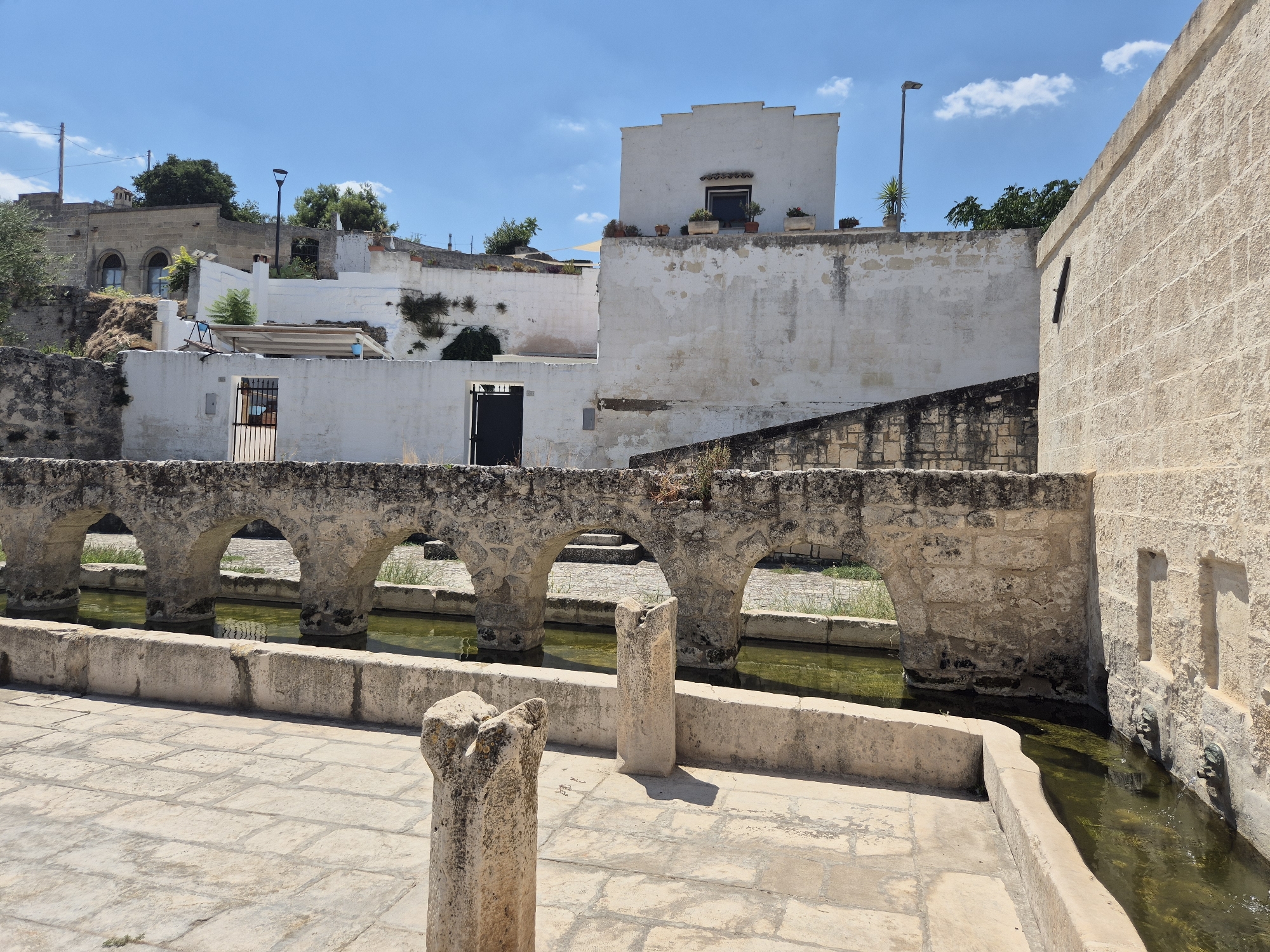 The image depicts an outdoor scene in Laterza, Italy, focusing on an ancient aqueduct structure with arched stone passageways. The aqueduct spans across a channel of murky green water. In the foreground, a paved courtyard is visible, with two weathered stone pillars. Beyond the aqueduct, white-washed buildings with flat roofs and some greenery are set against a clear blue sky with a few wispy clouds. The architecture suggests a historical Mediterranean setting. There are no people or animals visible in the image. The lighting indicates it is daytime, with the sun casting shadows.