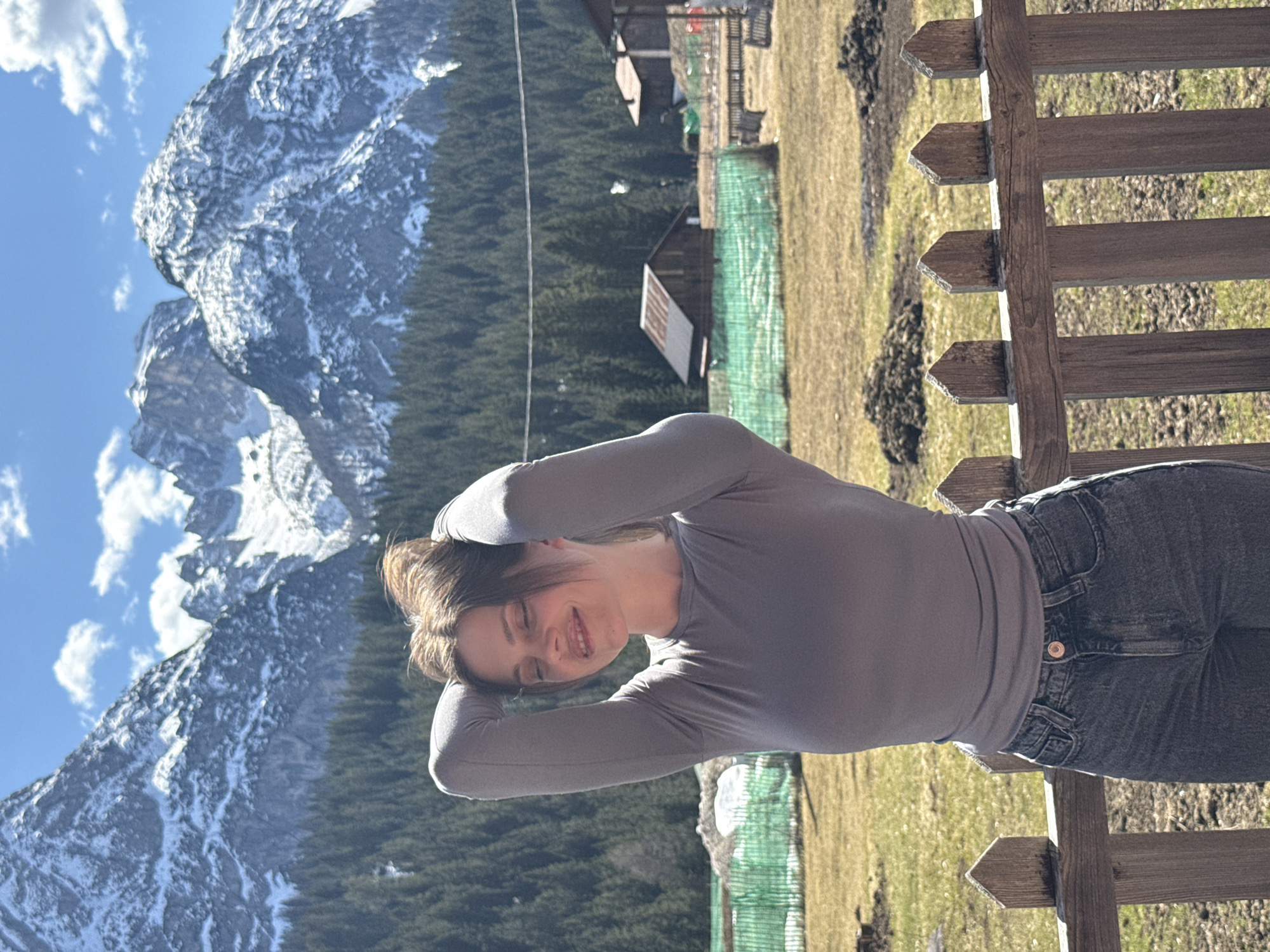 A medium shot presents a young woman with her hands behind her head, positioned in front of a weathered wooden fence. She is wearing a gray long-sleeved shirt and dark jeans. Beyond the fence stretches a landscape of grassy terrain dotted with what appears to be animal burrows. In the background, dense coniferous trees cover the hillside. In the distance, towering snow-capped mountains punctuate the scene, set against a blue sky with scattered clouds. The photograph was taken near Sappada / Plodn / Sapade, Italy.