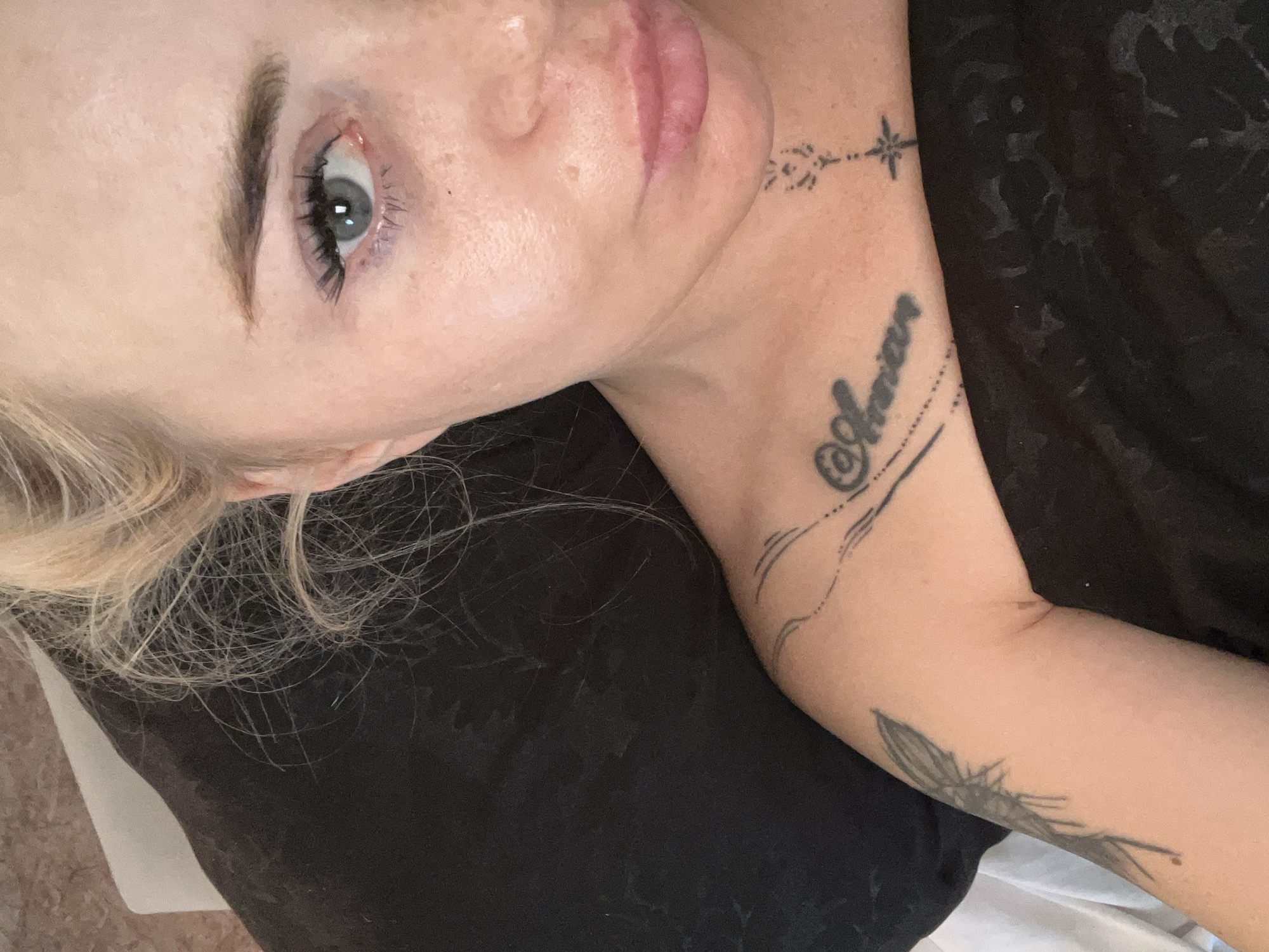 A close-up, over-the-shoulder selfie shows a woman lying down with her head on a dark pillow. She has blonde hair, blue eyes, and light skin with freckles. She is wearing a dark top, and several tattoos are visible on her arm and chest. One tattoo on her chest reads "@Adrian." Another tattoo on her arm appears to be a drawing of a dragonfly. The background is out of focus and appears to be a textured wall.