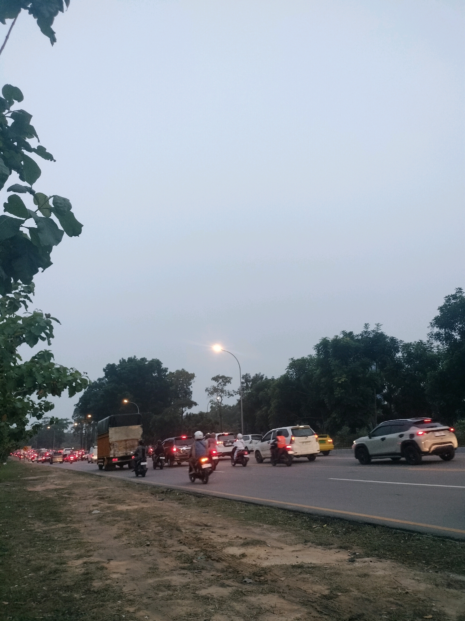 A dense traffic jam is depicted on a multi-lane asphalt road, possibly during twilight or overcast conditions given the light sky and artificial lighting from streetlamps. The scene appears to be in Batam, Indonesia, as suggested by the tropical foliage on the left and the style of vehicles, including motorcycles common in Southeast Asia.
On the road, numerous vehicles are visible, including cars, motorcycles, and a large truck with a covered cargo bed. The vehicles are stopped or moving slowly, indicating a significant traffic congestion. Several cars have their brake lights illuminated. Motorcycles are interspersed among the cars, with riders wearing helmets. A white car with off-road tires is prominent in the foreground on the right side of the road.
The environment consists of a grassy verge and dirt shoulder on the left side of the road, separating it from the traffic. Dense trees line the background on both sides of the road, with some appearing as thick foliage and others as taller, sparser trees. Streetlights are positioned along the road, casting a warm glow. The sky is a pale, overcast blue, suggesting a lack of direct sunlight. The overall impression is one of a busy urban or suburban road experiencing heavy traffic, likely in the late afternoon or early evening.