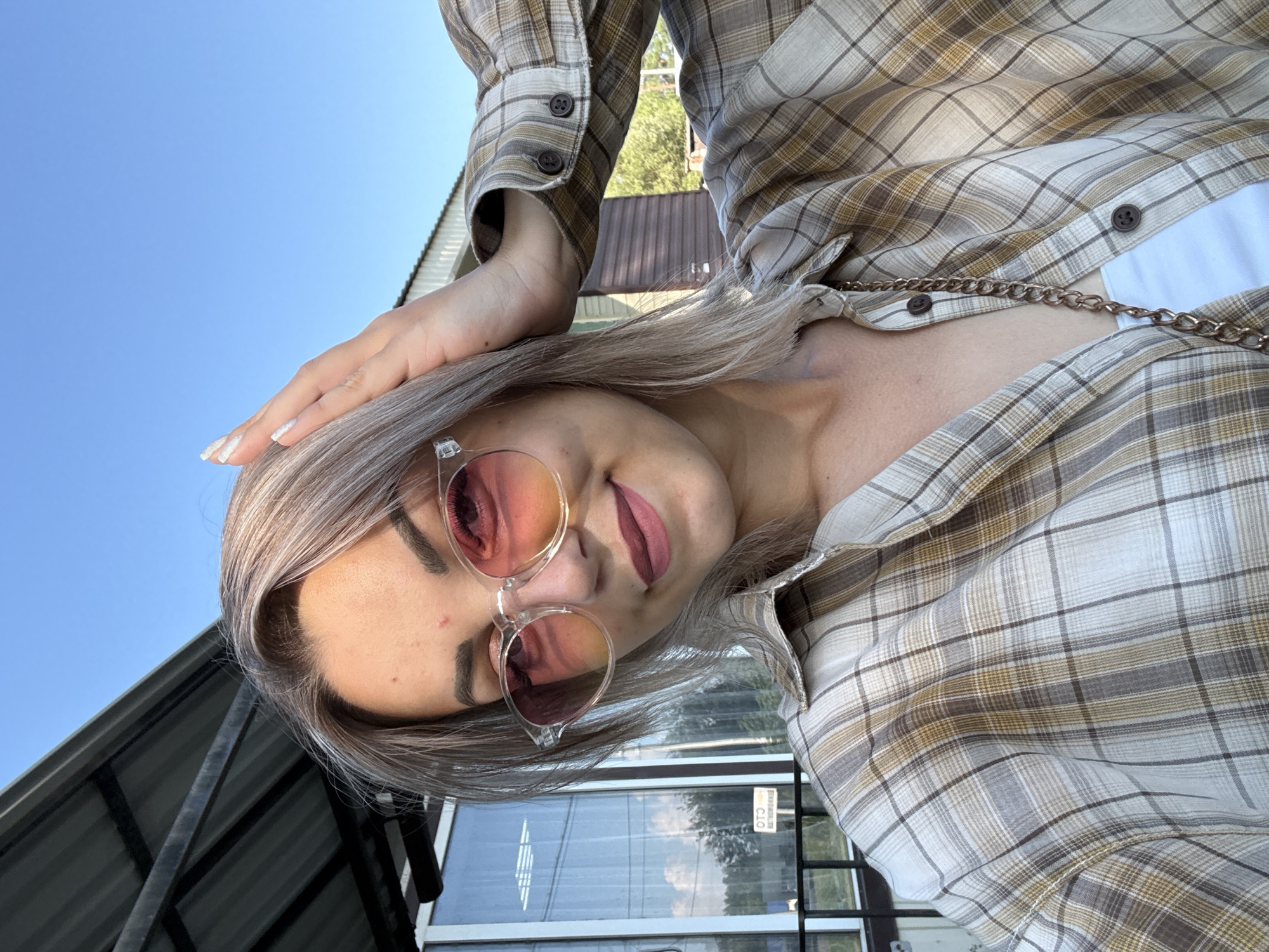 In the city of Krasnogorskoye, Russia, a young woman with blonde hair, wearing round, gradient pink-orange sunglasses and a plaid shirt over a white top, poses for a selfie outdoors. She is touching her head with her right hand. The background includes a blue sky, a building with a corrugated metal roof, and a glass-paneled structure with reflections of trees and clouds.