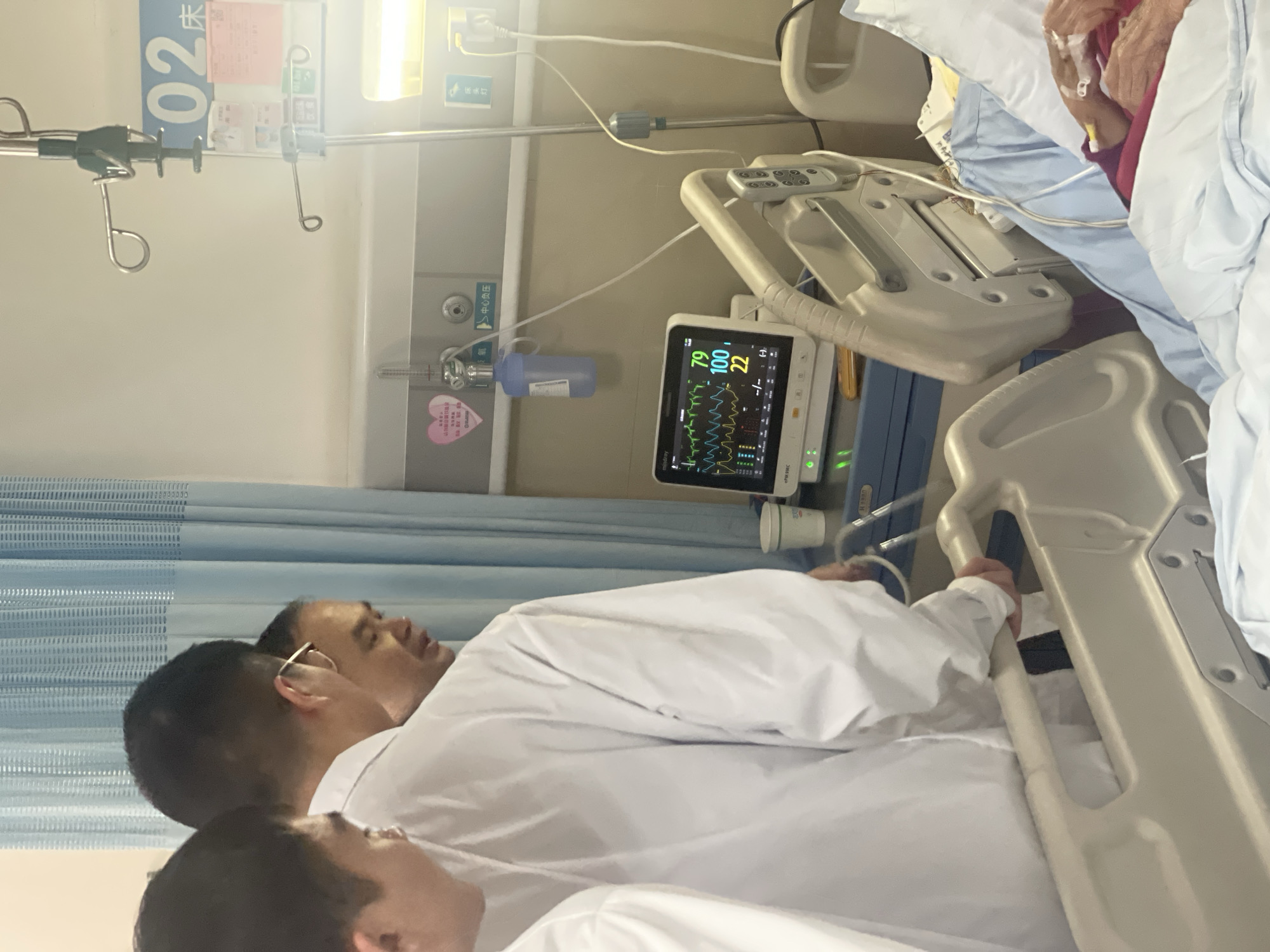 A hospital room in Tongliang District, China, features a patient in a bed with medical equipment. A cardiac monitor displays vital signs: heart rate (79), oxygen saturation (100), and respiratory rate (22). A medical professional in a white coat is present, with another person in a white coat partially visible in the lower left. An IV stand and other medical supplies are present in the room.