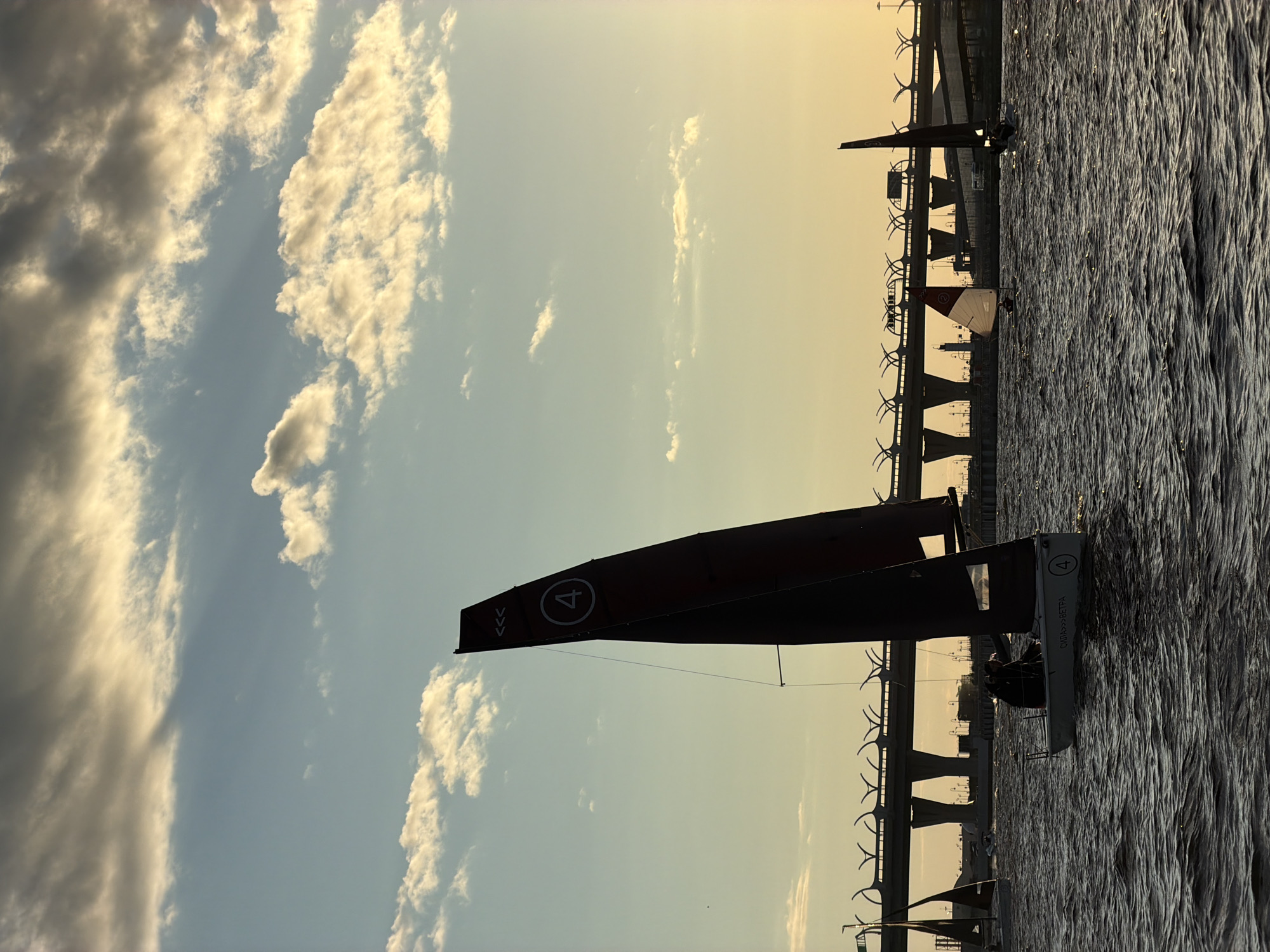 The image depicts a body of water, likely a river or bay, with sailboats navigating on it. A prominent feature is a large, modern bridge spanning the water in the background, with its distinctive curved support structures. The sky is partly cloudy, with sunlight filtering through, suggesting either sunrise or sunset. The overall atmosphere is serene, with the water rippling gently. In the foreground, one sailboat is closer to the viewer, its dark sail silhouetted against the sky, with a number "4" visible on it. Two people are seated in this sailboat. Further in the distance, other sailboats are visible, and the bridge extends across the horizon. The scene appears to be in Saint Petersburg, Russia, given the distinctive architecture of the bridge, which resembles the Western High-Speed Diameter. The image captures a moment of leisure or sport on the water, with the urban infrastructure of the city forming a backdrop.
