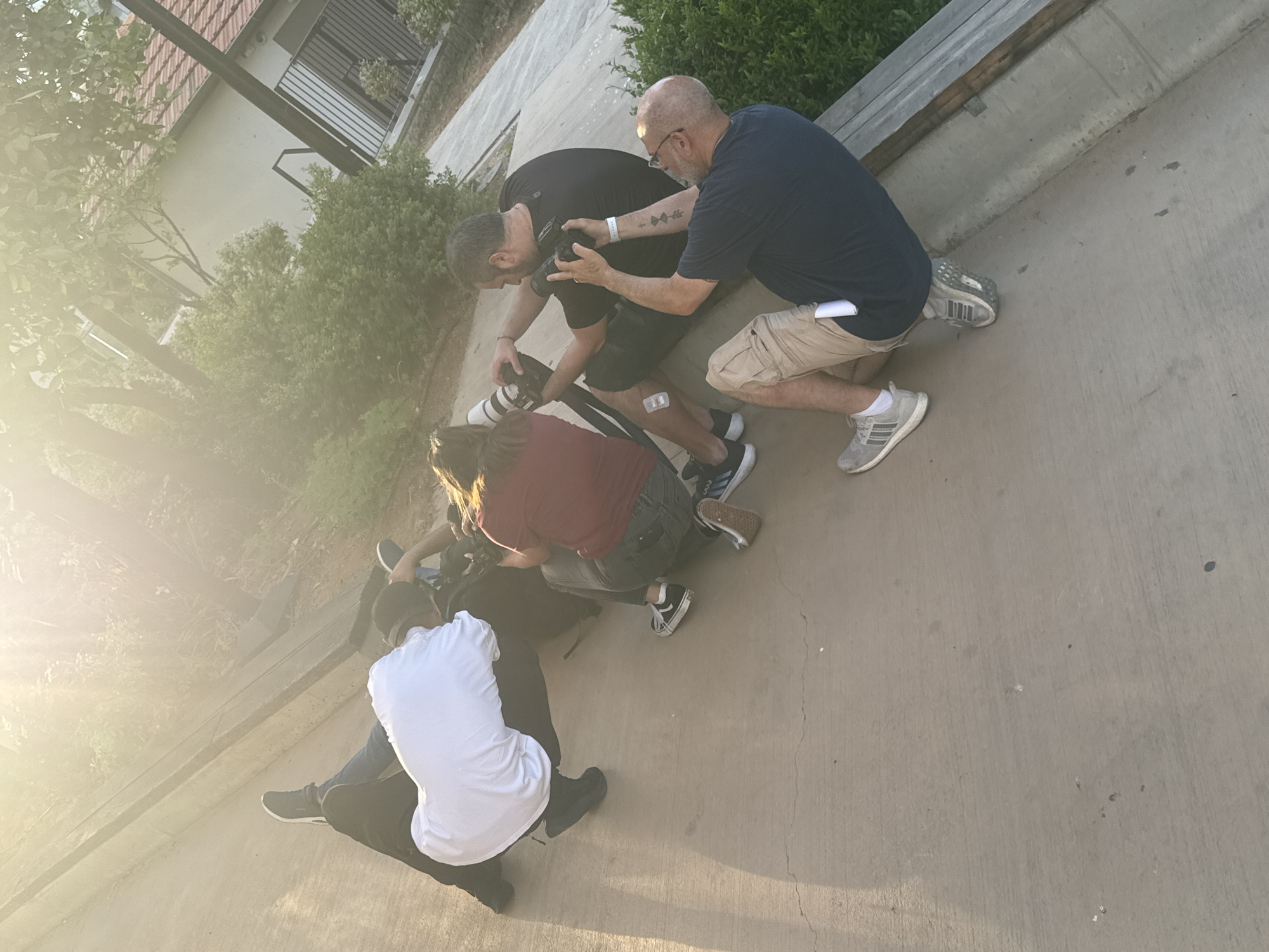 This media file captures an outdoor scene in what appears to be Tel-Aviv, Israel, showing a group of at least five individuals engaged in a photography activity. The image is taken from a slightly elevated, oblique angle, and bright lens flare from the sun illuminates the left side of the frame, indicating either early morning or late afternoon.
The primary subjects are two men in the foreground, kneeling and interacting with cameras. One man, with a shaved head and glasses, wearing a dark blue t-shirt and khaki shorts, kneels on his left knee while holding a camera. He appears to be instructing or demonstrating to another man beside him. This second man, with a beard and wearing a black t-shirt and black shorts, is also kneeling and holds a camera with a large, white-barreled lens, possibly a telephoto lens, pointed downwards. A small bandage is visible on his right knee. A white wristband is on the left arm of the man in the blue shirt, and a faint tattoo with geometric or arrow-like symbols is visible on his forearm.
Further to the left and partially obscured by light and other people, three more individuals are visible, also crouched low to the ground. One wears a white t-shirt, another a reddish-brown shirt. Multiple cameras and related equipment, including a tripod or monopod, are discernible among the group, reinforcing the photography context.
The setting is an outdoor concrete area, possibly a paved walkway or courtyard. Green foliage, including bushes and trees, frames the scene, with a building featuring a tiled roof and a black-railed balcony visible in the background, partially hidden by the greenery. A concrete ledge or bench runs along the right side of the frame. The strong, warm light creates visible shadows and a vibrant, focused atmosphere among the participants. No clearly legible text is visible on clothing or objects beyond the described tattoo design.