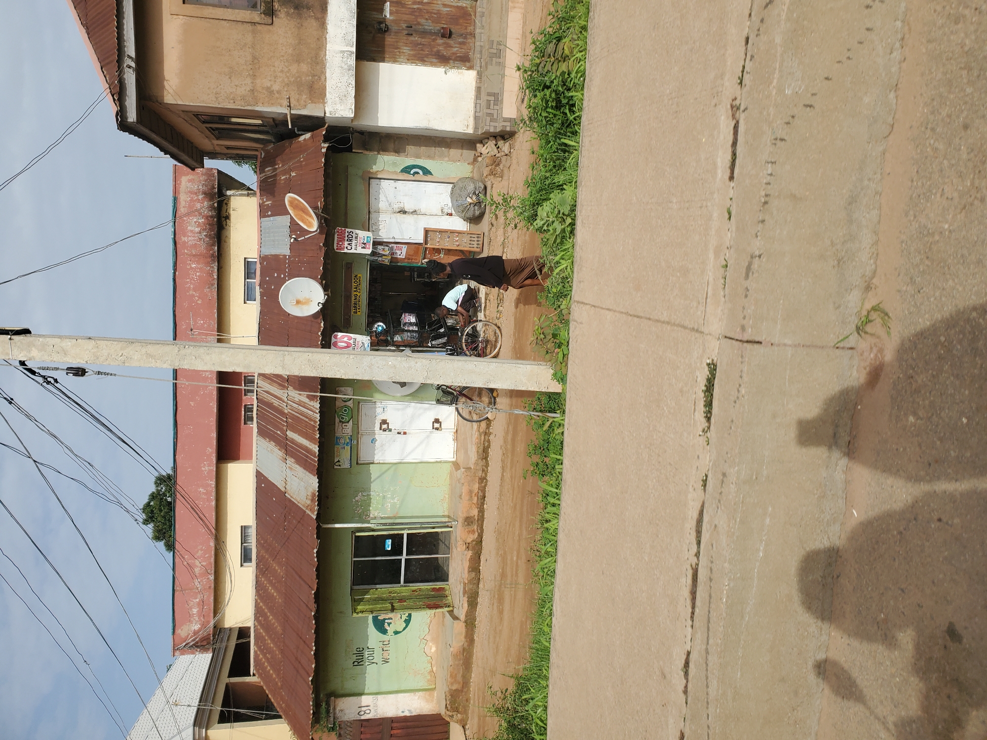The image captures a street scene in Bukuru, Nigeria. In the foreground, a paved surface, possibly a road or sidewalk, dominates the lower portion. The shadows of two individuals are cast upon this surface, suggesting they are positioned beyond the frame or are the source of the camera's perspective.
The mid-ground features a light green, single-story building with a corrugated metal roof. The building appears to house a business, with its storefront open. A sign visible on the building reads "BARBING SALOON & ELECTRICAL ELECTRONICS." Various items are displayed for sale within the open storefront, including what appears to be electronic equipment and possibly hair care products. Two satellite dishes are mounted on the roof of this building. To the right of the storefront, a bicycle is propped up. A man, dressed in a dark shirt and a skirt-like garment, walks away from the building towards the right side of the frame. Another individual is partially visible inside the storefront.
Behind this primary building, a taller, two-story structure with a reddish-brown, textured facade and a similar corrugated roof is visible. Further behind that, another building with a light yellow facade and a reddish-brown roof can be seen. The sky above is a pale blue with some scattered clouds and a network of overhead electrical wires. The environment appears somewhat rural or suburban, with vegetation growing along the edge of the paved area and in front of the buildings. The buildings show signs of wear and tear, typical of older structures.