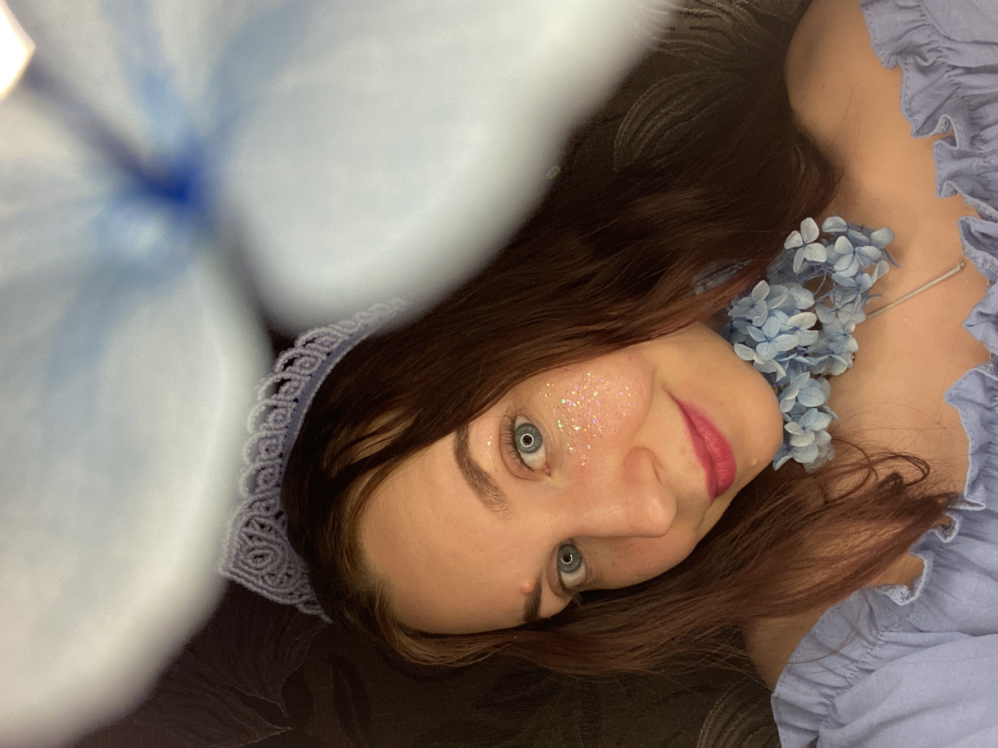 A woman is shown lying down in the image, with her head tilted to the side. Her hair is auburn and partially covers her face. She has blue eyes and is wearing makeup, including pink lipstick and glitter on her cheek. Around her neck, she is wearing a necklace of blue flowers that match the blue ruffled top she is wearing. Part of a large, blurred blue flower is in the foreground. The background is dark. No information is available in the image to identify the location as Chaikovsky, Russia.
