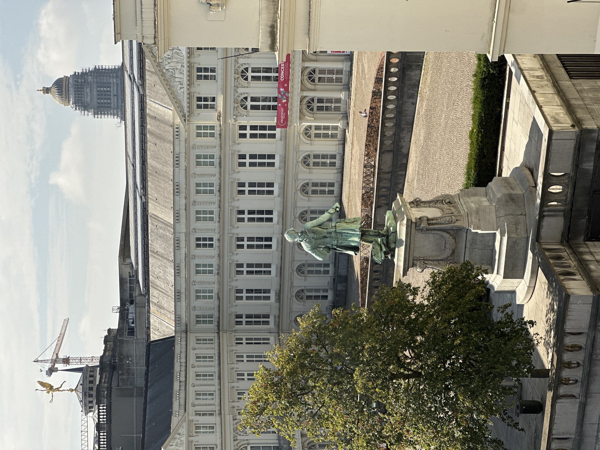 The image shows a view from an elevated position overlooking a courtyard in Brussels, Belgium. In the foreground, a bronze statue of a man stands on a stone pedestal. To the left of the statue is a large, leafy green tree. Behind the statue and tree is a large, ornate white building with many windows. A red banner with the text "musicorum CONCERT" is displayed on the building. The courtyard is paved with cobblestones. In the background, a towering building with a domed roof, possibly under construction, is visible, along with a construction crane and a golden statue on top of another building. The sky is partly cloudy. A single person is visible in the courtyard, standing near the building.