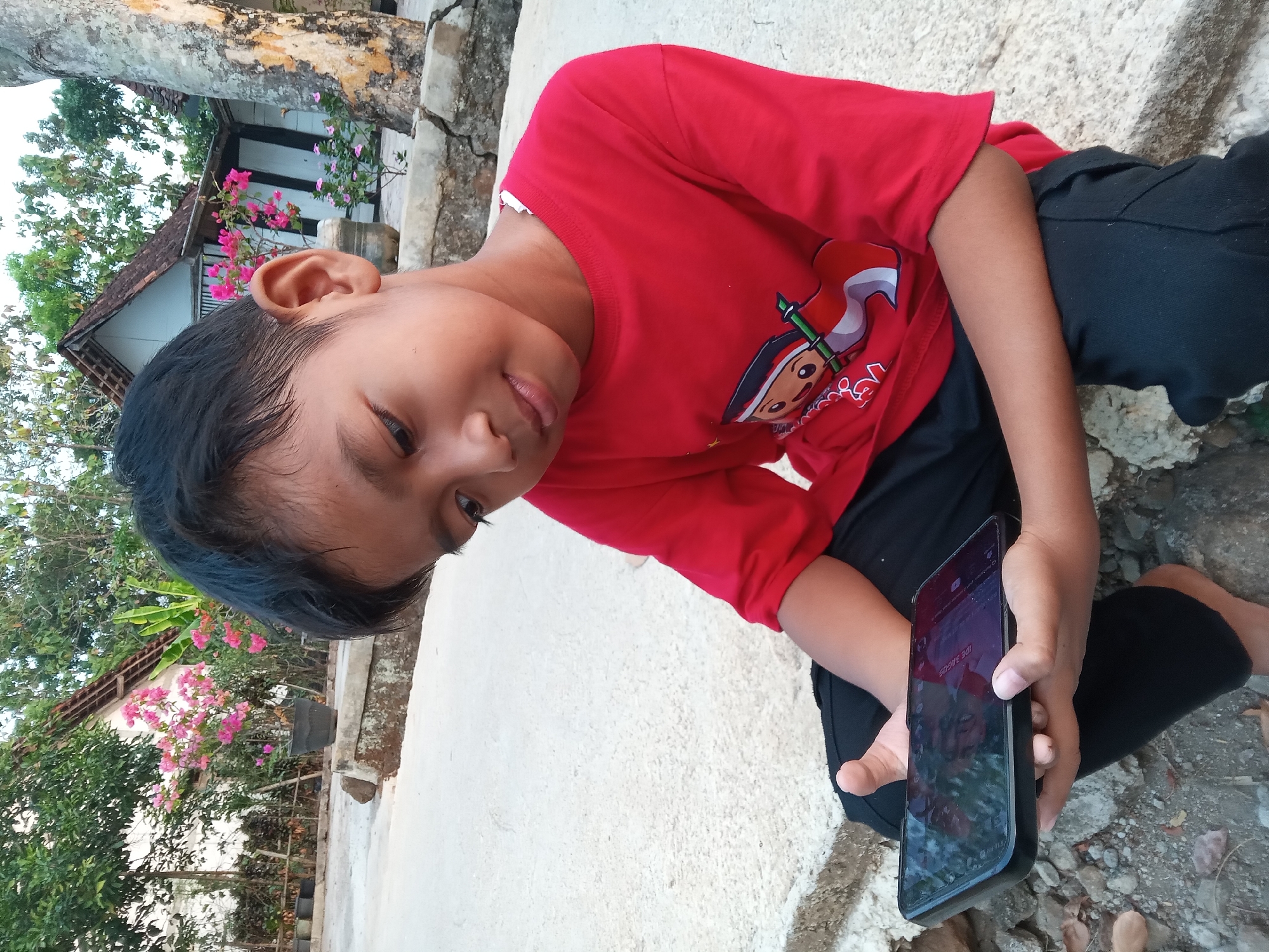 A young boy in a red long-sleeved shirt and black pants sits on a concrete surface outdoors, looking down at a smartphone he holds in his hands. He is positioned in the foreground, with his body angled slightly to his right. The phone's screen is illuminated, displaying some content. In the background, there are two traditional Indonesian houses with tiled roofs and white walls. Pink flowering plants in pots are visible near the houses, adding a touch of color to the scene. A large tree trunk stands to the right of the frame, and the ground is a mix of concrete and loose gravel. The overall setting appears to be a residential area in the village of Kedunggudel, Indonesia, suggesting a casual, everyday moment.