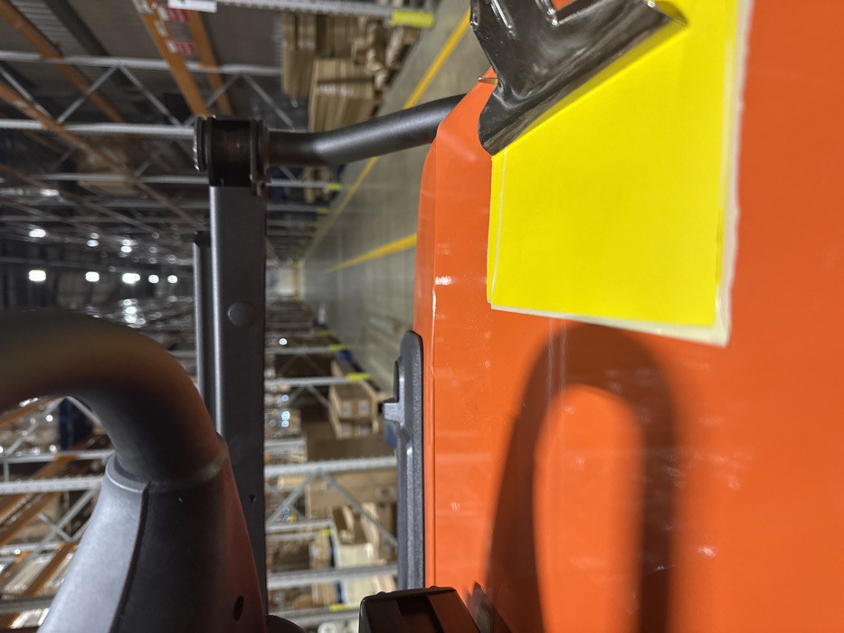 This image depicts the interior of a large industrial warehouse or distribution center, located in Kettering, United Kingdom. The perspective is from the operator's position of heavy machinery, likely a forklift or order picker, looking down a lengthy aisle.
In the immediate foreground, an orange, glossy surface, consistent with a vehicle's console or body panel, occupies the lower frame. On this surface, a bright yellow rectangular sheet of paper or card is secured on the lower right by a reflective, silver-colored metal clip. A dark, curved shadow is cast on the orange surface from the left.
Above the orange surface, dark gray tubular and structural components of the machinery's frame are visible, partially obscuring the view into the aisle.
The background showcases an extensive warehouse environment with tall, multi-tiered metal racking systems lining both sides of a wide aisle. These racks hold numerous palletized goods and cardboard boxes. The concrete floor features prominent yellow demarcation lines, indicating a clear pathway that recedes into the distance. Overhead industrial lighting fixtures are visible as bright, diffused sources of illumination. No people are present within the scene.