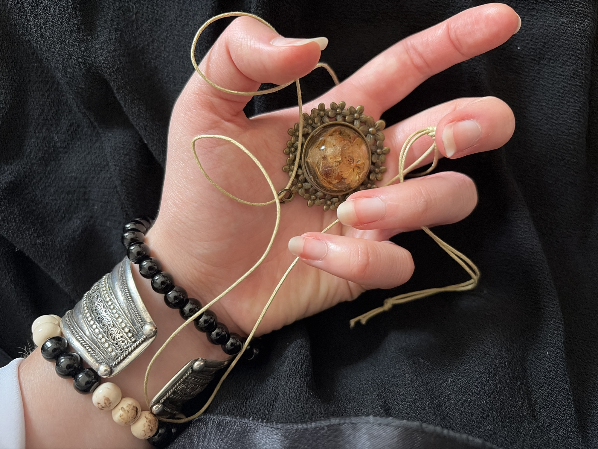 The image shows a person's hand wearing multiple bracelets and holding a necklace. The bracelets are made of black beads and carved bone or stone beads, with decorative silver accents. The necklace has a pendant with a round, amber-colored inclusion surrounded by a floral design. The hand is resting on a black fabric. The context of Chelyabinsk, Russia, is not present in the image.