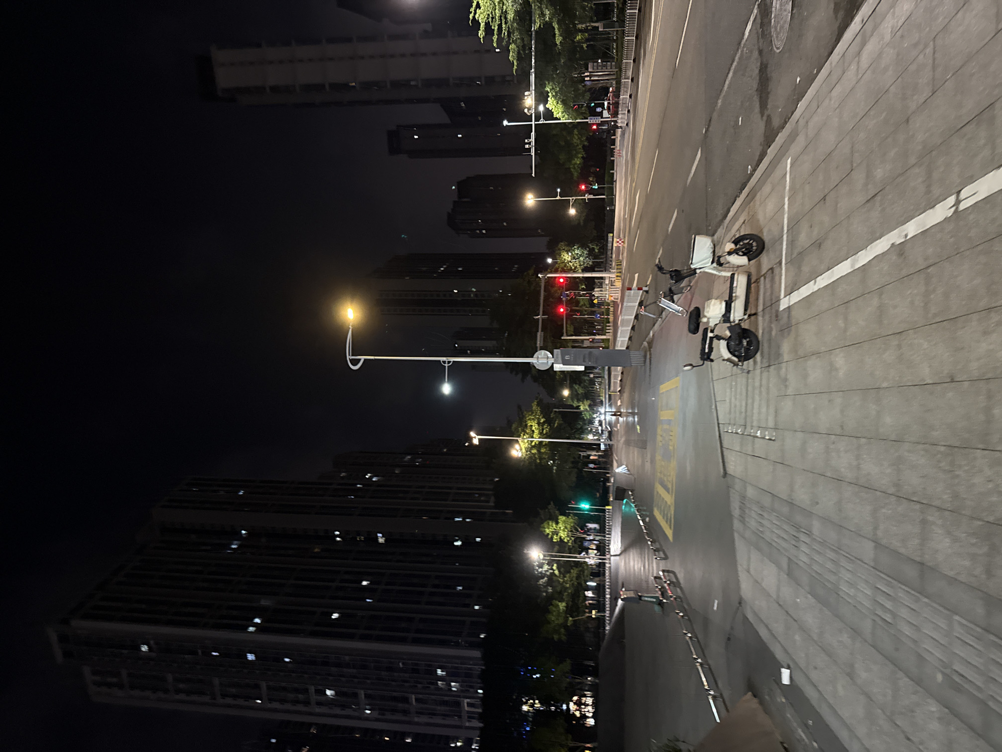 The image depicts a street scene at night in Haizhu District, China. The foreground features a pedestrian walkway paved with grey rectangular tiles, with tactile paving elements visible. A light-colored electric scooter is parked on the walkway. Adjacent to the walkway is a road with multiple lanes, marked by white lines. Traffic lights are visible, some showing red. Tall residential or office buildings with illuminated windows line the street on the left and in the background. Trees are present along the sidewalk and between buildings. Streetlights cast an orange glow on the scene. The sky is dark and clear.
