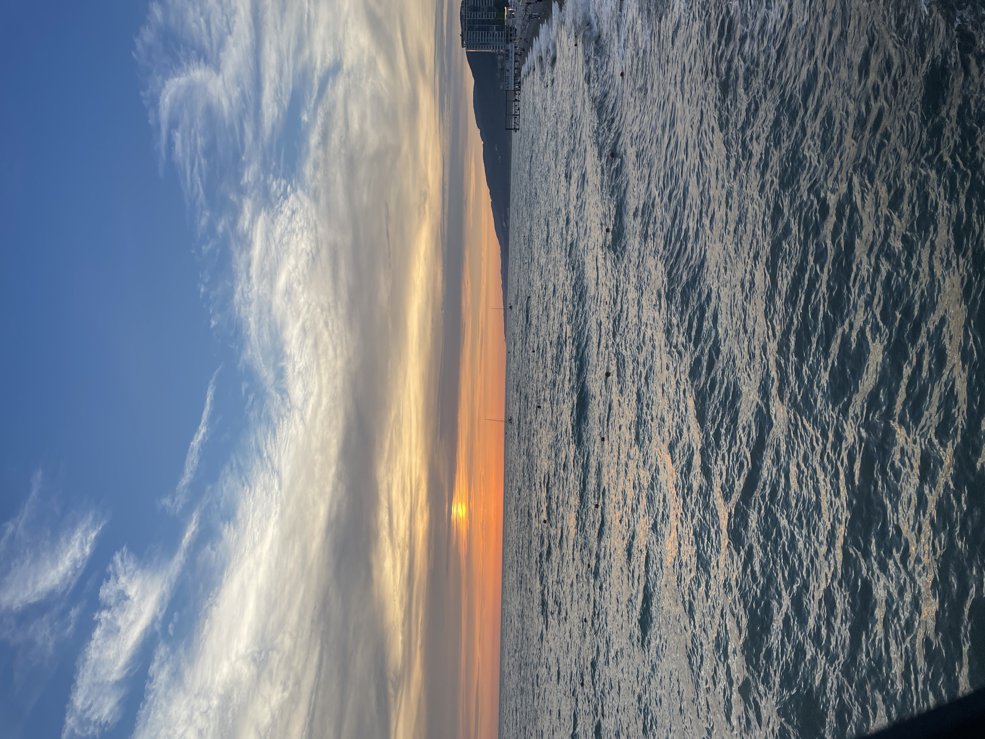 A wide-angle, eye-level, outdoor shot depicts a sunset over a calm sea with a beach in the background. The sky is a gradient of deep blue at the top, transitioning to orange and yellow hues near the horizon. Wispy white clouds are scattered across the sky. The sun, a bright orange circle, is visible just above the horizon line. The sea is a deep blue with gentle waves creating ripples on the surface. Small red buoys are dotted across the water. On the right side of the image, a sandy beach is visible, lined with a few palm trees and a pier extending into the water. Several buildings, including a tall, modern hotel, are situated behind the beach. The overall scene is serene and peaceful, capturing the beauty of a coastal sunset.