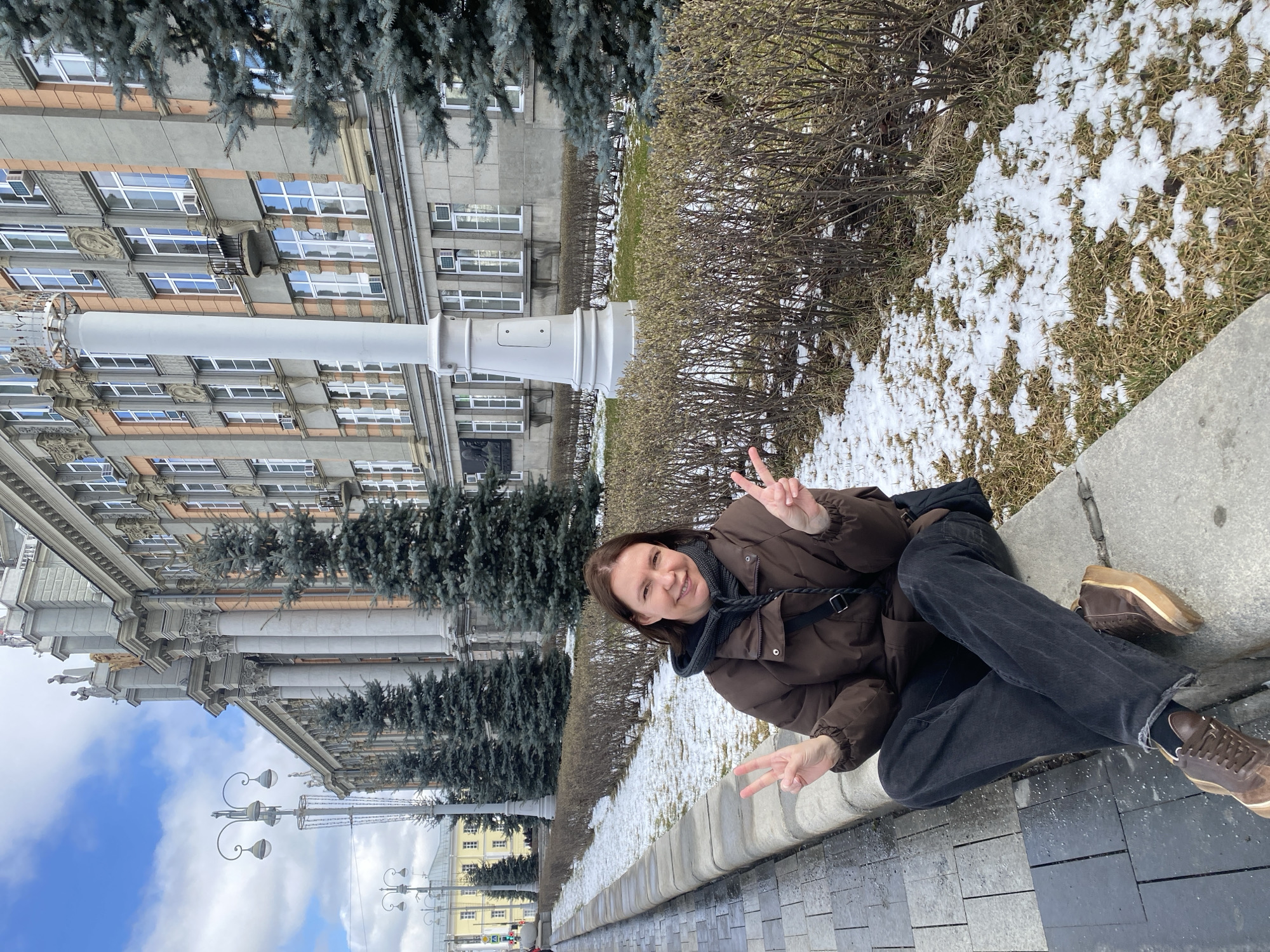 A woman is seated on a concrete ledge with a building and landscaping in the background. The woman is wearing a brown puffer jacket, a dark scarf, dark jeans, and brown shoes. She is making a peace sign with both hands and smiling at the camera. Behind her is a hedge with patches of snow. The background features a large building with multiple windows and columns, flanked by evergreen trees. Streetlights and other buildings are visible in the distance. The location is Yekaterinburg, Russia. The photograph is rotated clockwise 90 degrees.