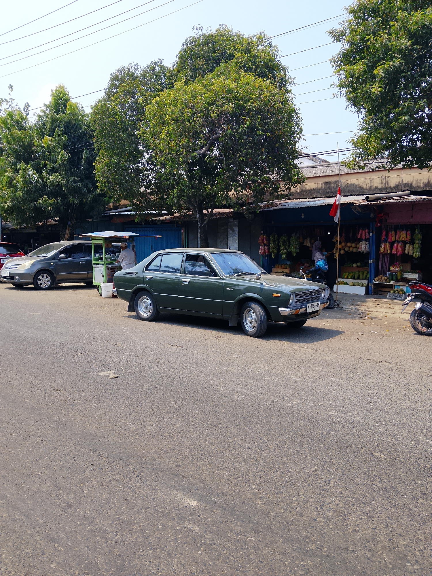 In the city of Ngelo, Indonesia, a green sedan is parked on a paved road next to a street vendor's cart. The vendor, a person wearing a light-colored uniform, is serving a customer. Behind the vendor's cart, there's a shop selling various produce, with a red and white Indonesian flag displayed prominently. Another car, a dark gray sedan, is parked further down the road. In the foreground, the asphalt road is visible, with some leaves and debris scattered on it. The background features trees and overhead power lines, indicating a sunny day.