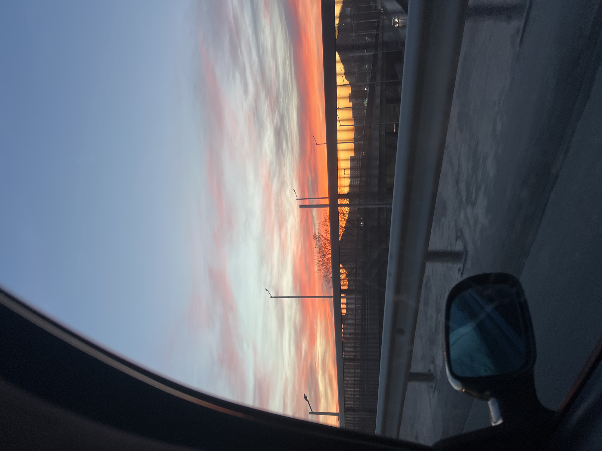 The image shows a view from inside a car, likely on a bridge or elevated roadway in Plovdiv, Bulgaria. The sunset is visible in the sky, with orange and pink hues. The railing of the bridge is visible on the right side, along with a car's side mirror. Streetlights and building silhouettes are seen in the background beyond the railing. The inside of the car is dark.
