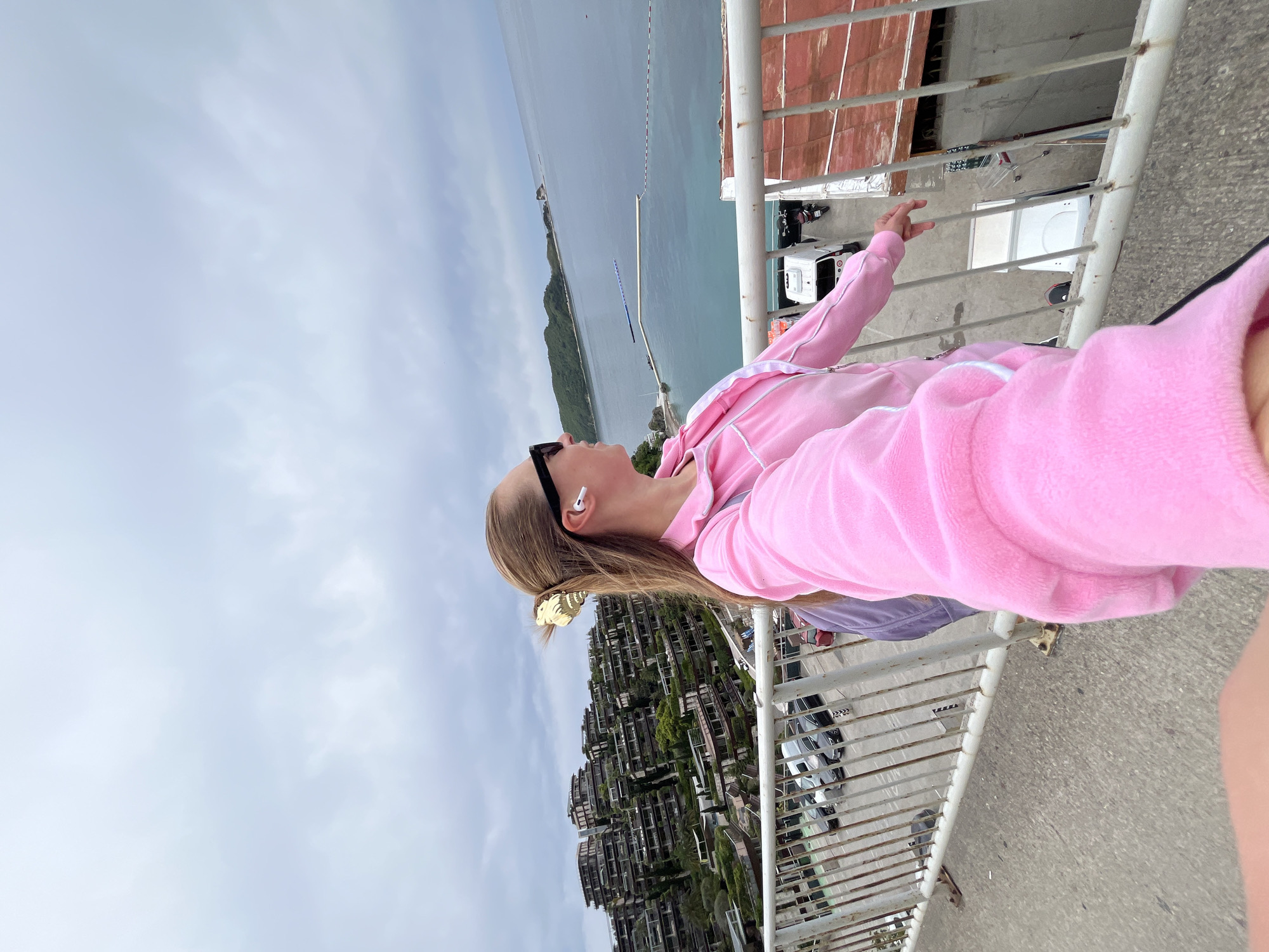 A medium shot captures a fair-skinned woman with long blonde hair wearing sunglasses, an earbud, and a pink hoodie, leaning against a metal railing, looking upwards. The background features a body of water, potentially the Adriatic Sea, stretching into the horizon with a strip of land visible in the distance. To the woman's left, multiple buildings cascade down a hillside, typical of urban development in mountainous coastal regions. A section of pavement is visible below the railing, indicating a pedestrian area. The image's perspective suggests it was taken from an elevated position, likely a bridge or high vantage point. The location appears to be in or around the city of Budva, Montenegro, based on the architectural style and coastal landscape. The focus is on the woman, but the urban and natural environment provides significant context.