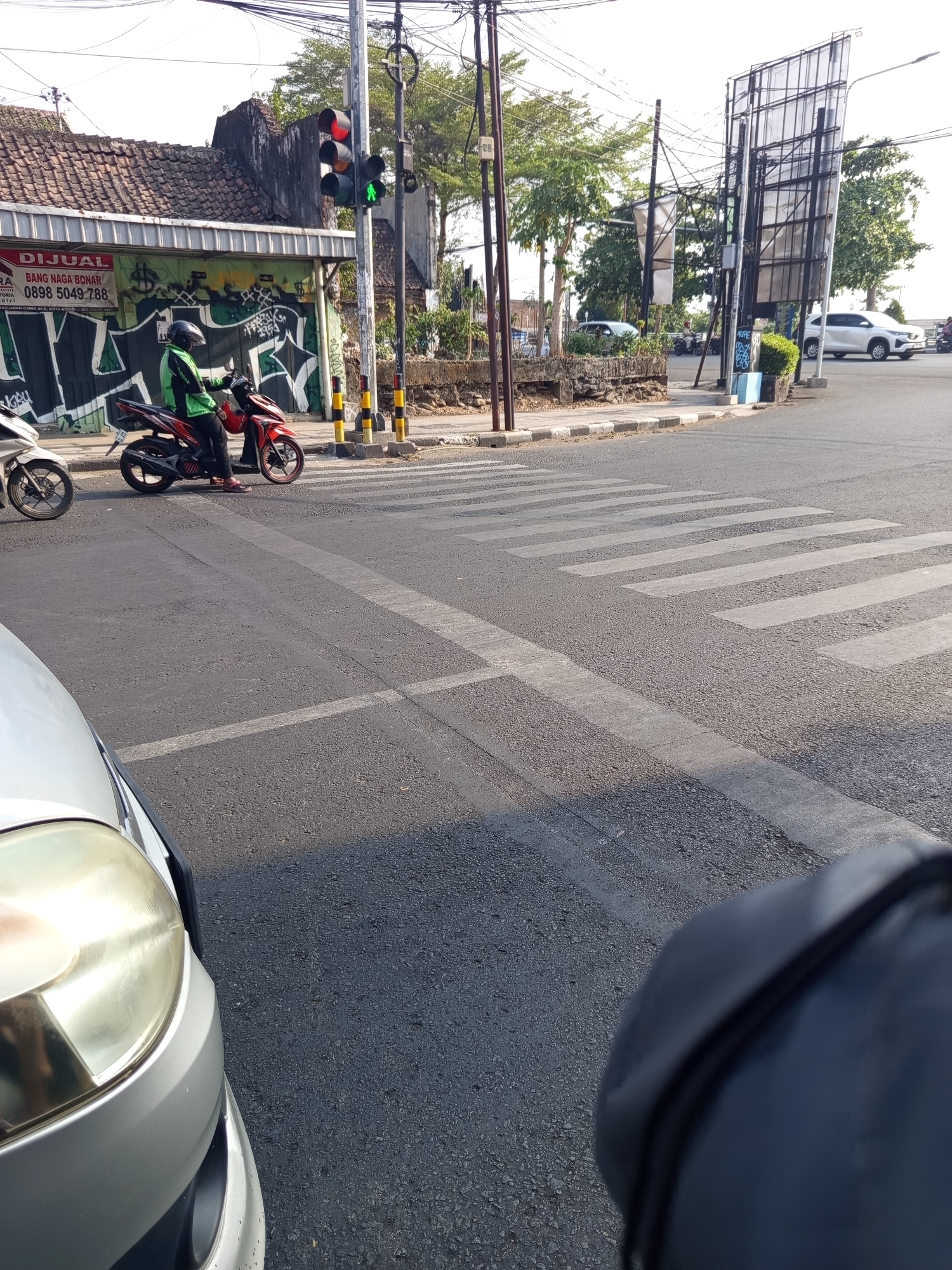 The image depicts a street scene at a pedestrian crossing in Kediri City, Indonesia. A red motorcycle with a rider wearing a green jacket and helmet is stopped at the crossing, facing left. The rider's feet are on the ground. To the left of the motorcycle, another motorcycle is partially visible. The pedestrian crossing is marked with white stripes on the asphalt road. In the background, there is a building with a tiled roof and a sign that reads "DIJUAL BANG NAGA BONAR". A traffic light is visible, showing a red signal for vehicles. The environment includes trees, power lines, and a large billboard structure. A silver car is visible in the foreground, partially obscuring the view of the road.