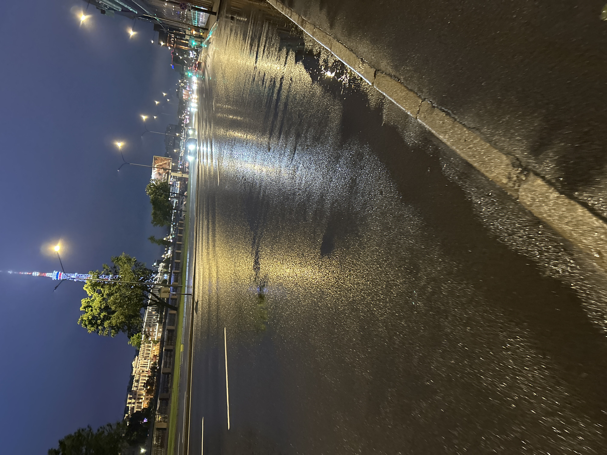 An eye-level shot captures a wet asphalt street at night, reflecting the artificial lights from street lamps and passing vehicles. The sky is a deep, dark blue, suggesting either twilight or night. In the background, a tall, brightly lit tower stands prominently, its structure illuminated with red, white, and blue lights. Adjacent to the tower, a row of buildings lines the waterfront, their windows glowing. Cars are visible on the road, their headlights and taillights creating streaks of light on the wet surface. The scene depicts an urban environment at night, with the street wet from recent rain or other precipitation.