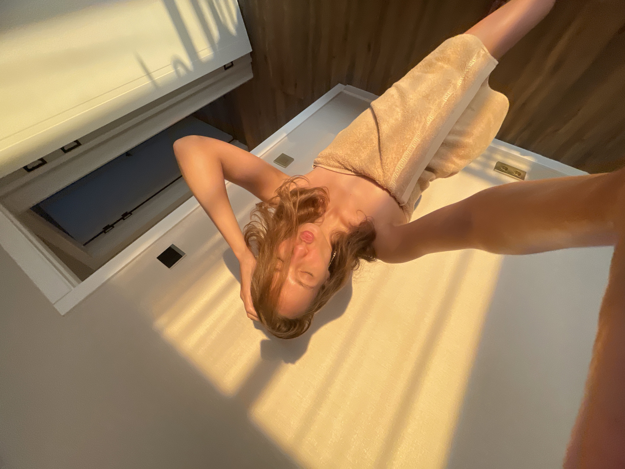 A woman, draped in a beige towel, poses for a selfie in a room. She has her eyes closed, lips puckered, and her left hand is placed on her head. Sunlight casts dramatic shadows from blinds across the wall and floor. The room appears to be a domestic interior, possibly a bedroom or bathroom, with doorways and wooden flooring visible. The location is specified as Podshivalovo, Russia.