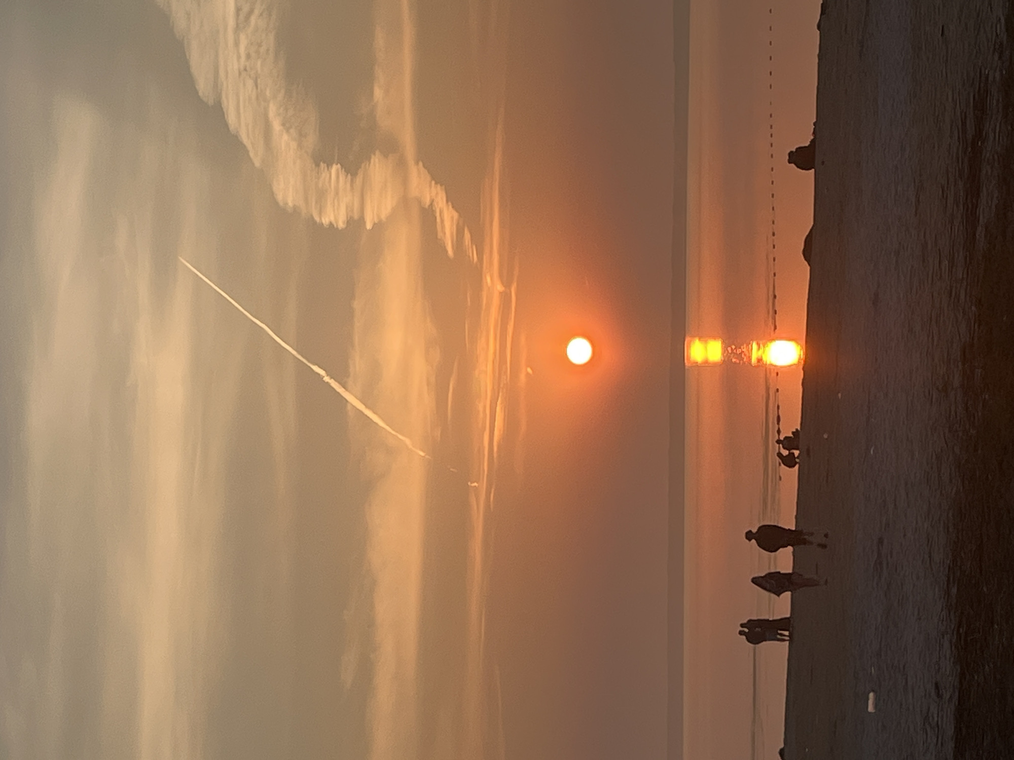 The image depicts a serene sunset over a body of water, likely the Volga River, as viewed from the shore in Surakovo, Russia. The sky is painted in hues of orange and yellow, with wispy clouds and a prominent contrail from an aircraft streaking across the upper portion. The sun, a bright orb, is partially submerged on the horizon, casting a shimmering reflection on the calm water. On the sandy beach in the foreground, several silhouetted figures are visible. Some are walking along the shore, while others appear to be sitting or standing in small groups, enjoying the twilight. The scene is characterized by a peaceful ambiance, with the natural beauty of the sunset dominating the composition.
