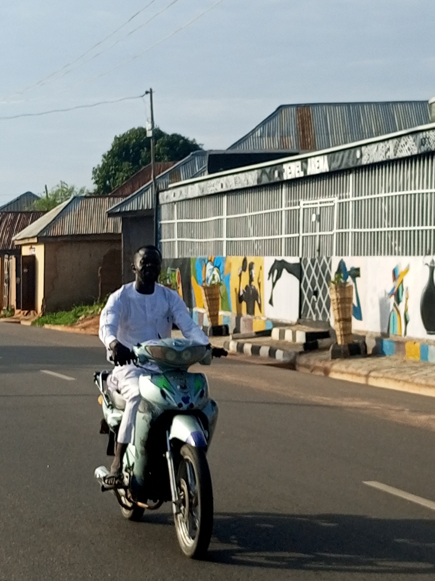 A man wearing a white traditional outfit rides a light blue and white motorcycle on a paved road in Gombe, Nigeria. The man is facing forward, and his head is turned slightly towards the viewer. He is wearing black gloves. The motorcycle has a large headlight and a windshield. In the background, there are buildings with corrugated metal roofs. One building has a mural on its wall depicting figures and abstract shapes. There is a sidewalk with a black and white striped pattern in front of the building with the mural. The sky is clear and blue.