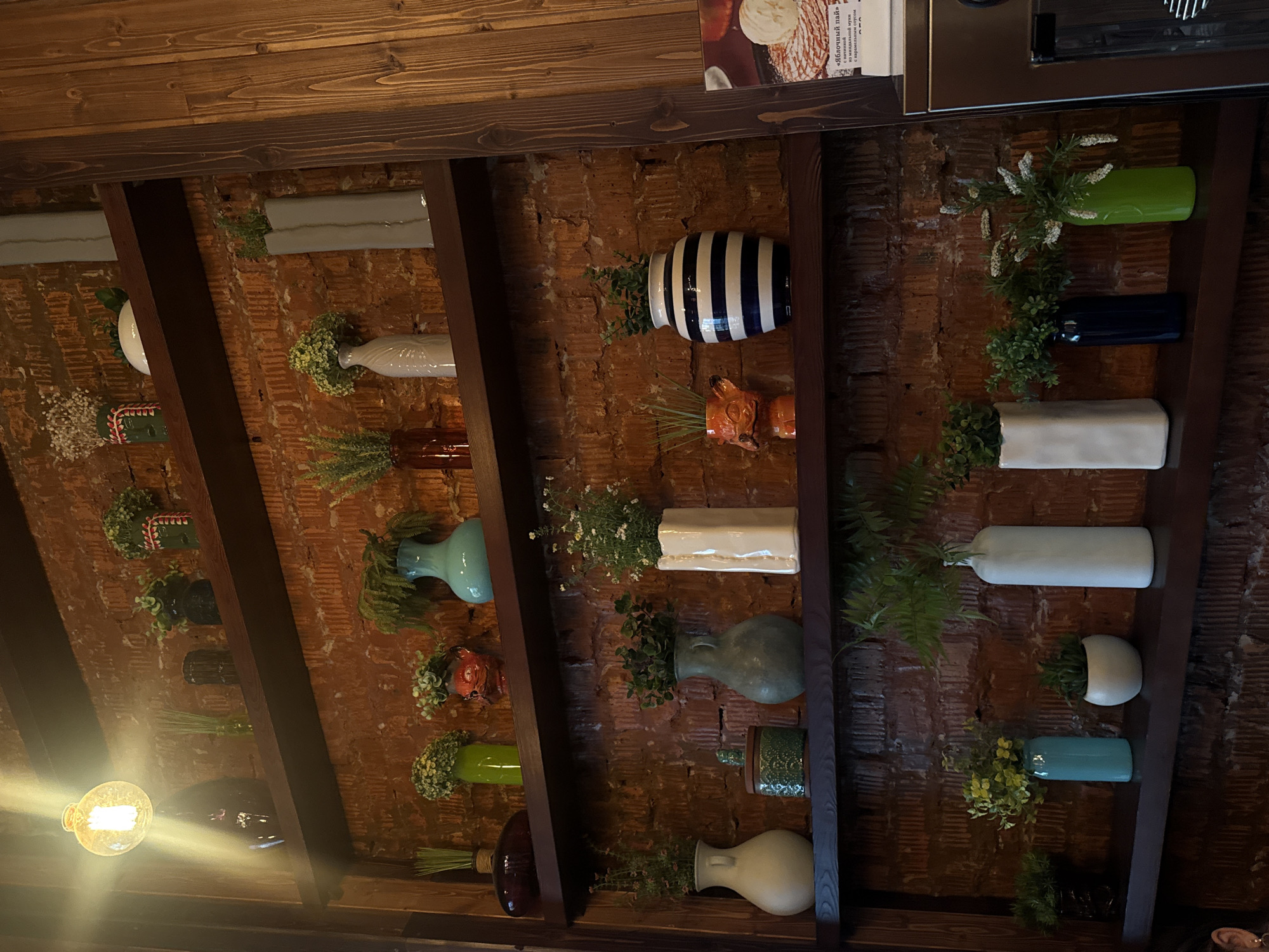 The image captures an indoor decorative wall, likely in a cafe or restaurant in Birobidzhan, Russia, viewed from a low angle looking upwards. The background features a rustic red brick wall, partially covered by a grid of dark wooden beams and slats that create shelving or a display structure.
Numerous ceramic and glass vases and planters, holding artificial plants and flowers, are arranged on or within this wooden framework, affixed to the brick wall. These decorative items come in a variety of shapes, sizes, and colors, including light blue, green, white, cream, dark blue, brown, grey, and one distinctive planter with blue and white stripes. The artificial greenery includes ferns, small white cluster flowers, and various leafy green plants, creating a lush, natural aesthetic against the brick and wood.
In the bottom left, a prominent, exposed filament light bulb glows brightly, casting a warm light across the scene. At the top right, a portion of a menu or flyer is visible, displaying an image of a dessert with ice cream and some text. Visible Russian text includes "Яблочный пай" (Apple Pie) and smaller text below that likely describes the item, such as "сладкий яблочный пирог" (sweet apple pie) and "подается с шариком мороженого" (served with a scoop of ice cream). Adjacent to the menu is a metallic surface, possibly part of a kitchen appliance or counter.
The overall impression is one of a cozy, rustic, and thoughtfully decorated space.