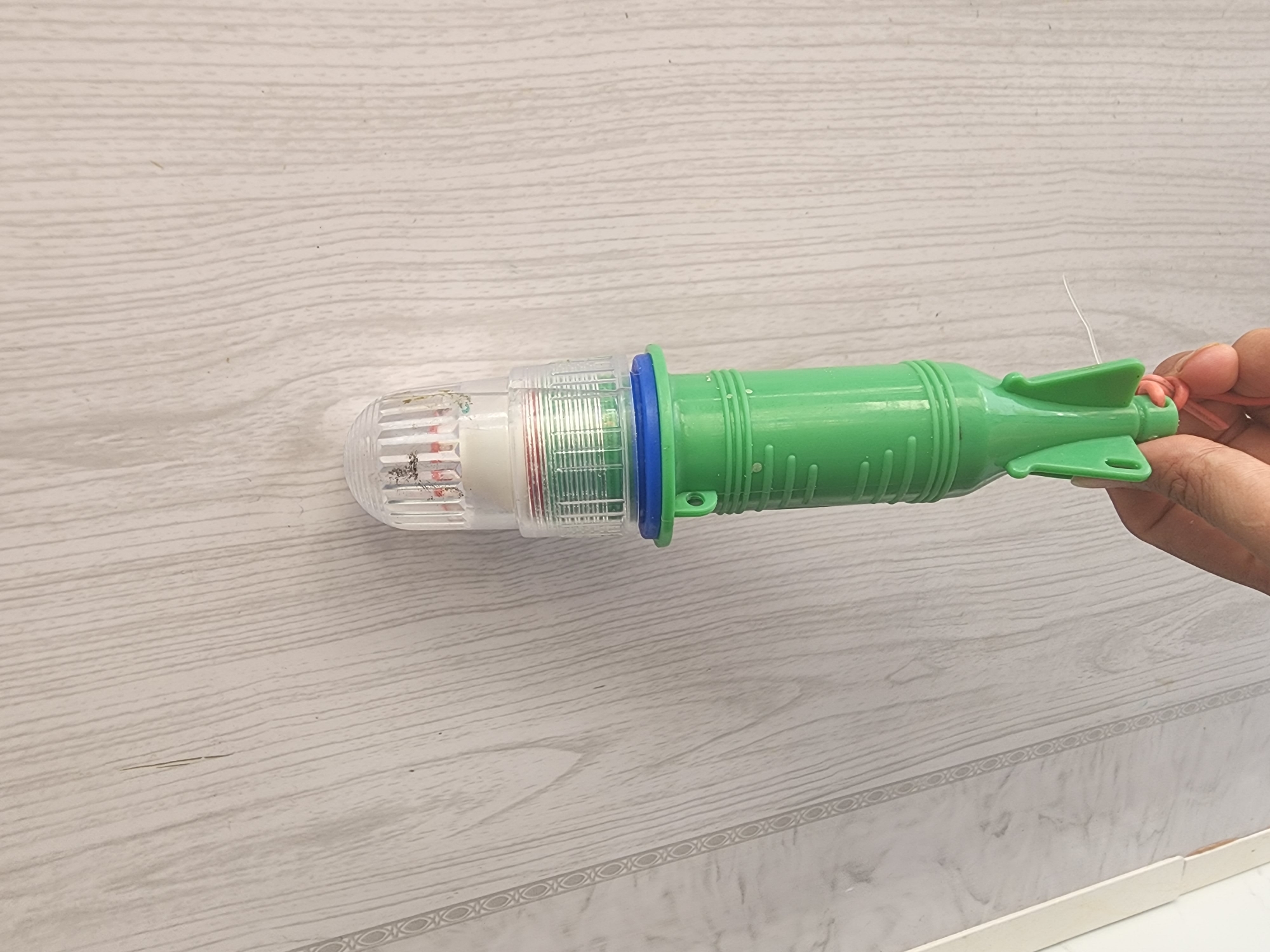 A hand is holding a green, rocket-shaped fishing light with a clear plastic top that contains red and white components and a blue ring around its base. The fishing light is positioned vertically against a light-colored, wood-paneled wall. The wall has a subtle, repeating decorative border of circular shapes. The lighting in the scene is neutral. The location is Phan Thiet Ward, Vietnam.