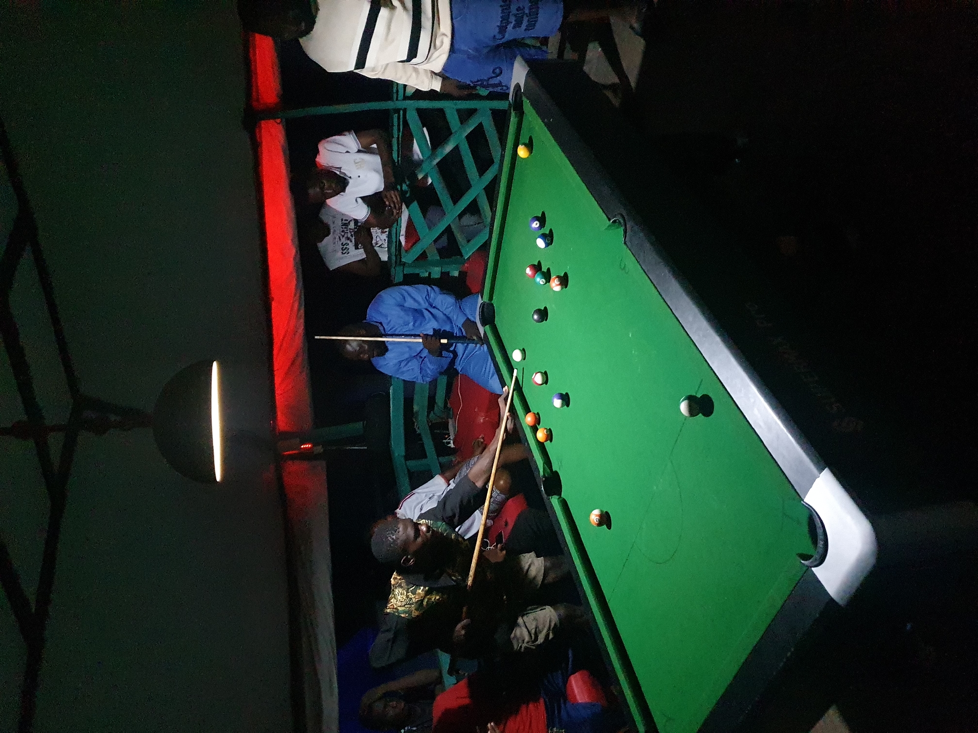 The image captures a nighttime scene at a pool hall, possibly outdoors or in a partially enclosed space, in Lokoja, Nigeria.
A pool table, covered in green felt and populated with pool balls, dominates the right side of the frame. A man in a blue shirt stands at the head of the table, cue in hand, poised to strike the cue ball. A few people, some seated and some standing, watch the game.
The backdrop includes a wooden or metal fence with a red light fixture illuminating the scene. A hanging light fixture provides additional illumination.