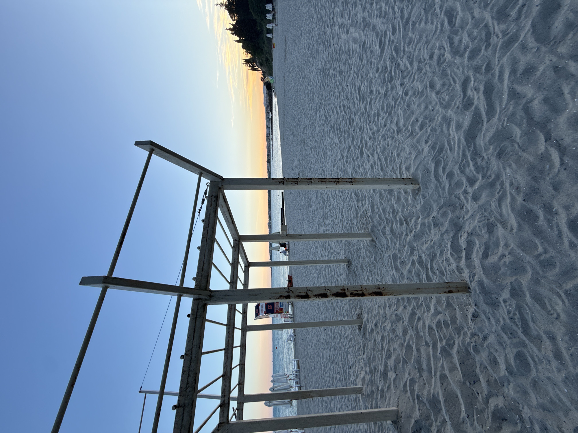 The image depicts a serene beach scene in Nesebar, Bulgaria, during the late afternoon or early evening. The foreground is dominated by a sandy beach with visible sand ripples, leading to the sea in the background. A white, weathered wooden structure, possibly a shelter or part of a playground, stands prominently in the left half of the frame, with its posts anchoring into the sand.
In the mid-ground, the sea is calm and reflects the soft colors of the sunset. Along the shoreline, several white beach umbrellas and lounge chairs are visible, suggesting a public beach area. A red rescue buoy is placed on the sand near the water's edge. A prominent advertising billboard for "Betano" is situated behind the wooden structure. Further down the beach, a few distant figures can be seen, including two individuals sitting on chairs and others standing near the water. The coastline extends into the distance, lined with buildings and lush green trees on a gentle slope. The sky is a clear gradient of blue, transitioning to hues of orange and yellow near the horizon, indicative of a sunset.