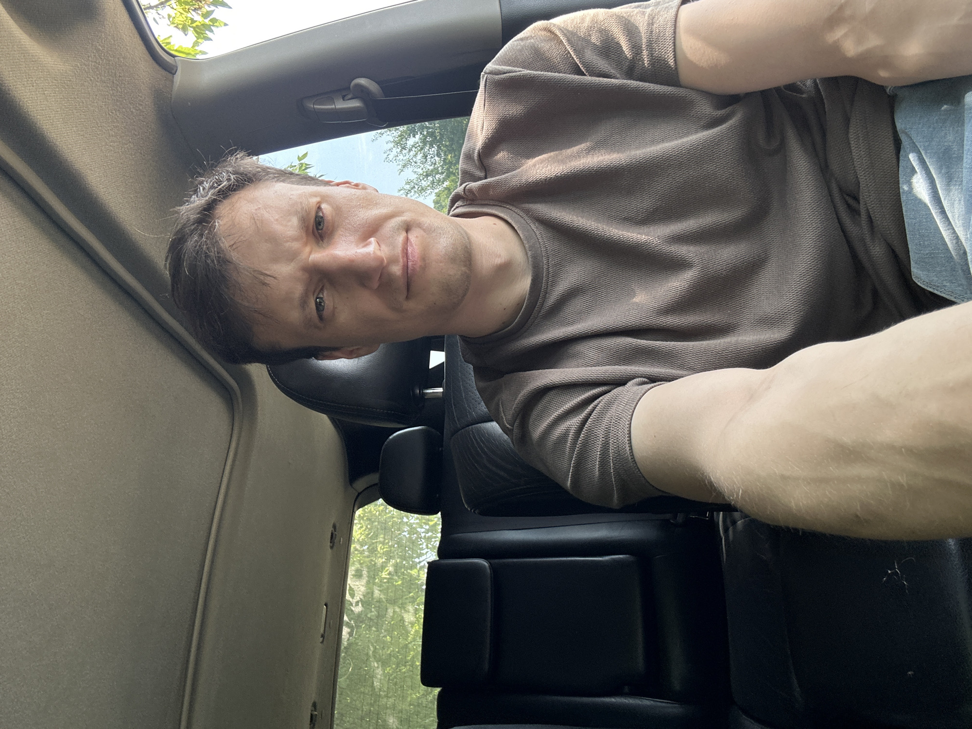 A selfie shows a man in his late 30s with short, dark brown hair, a light complexion, and blue eyes, looking directly at the camera with a neutral expression. He is wearing a brown, short-sleeved collared shirt. The man is seated in the front passenger seat of a vehicle, with the headrest and part of the car's interior visible. The background includes the back seat of the car, a sunroof overhead, and foliage visible through the windows. The image appears to have been taken outdoors, possibly in Makiv, Ukraine, given the natural light and greenery.