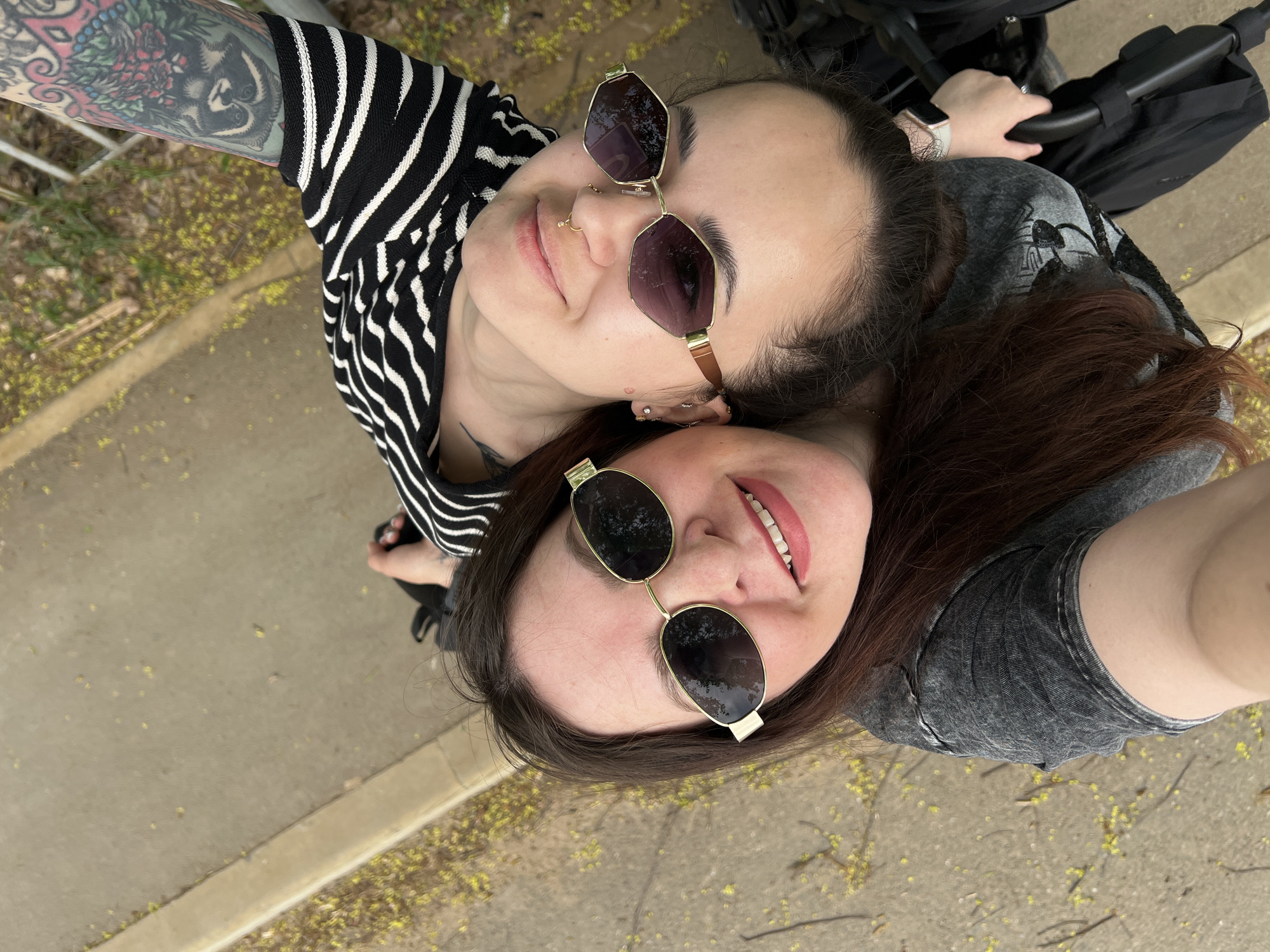 Two women are closely positioned, taking a selfie outdoors on a concrete surface with scattered yellow flowers. One woman is wearing a black and white striped shirt and sunglasses with a distinctive angular frame. Her left arm, visible in the upper left of the frame, is covered in tattoos. She has a nose ring and multiple piercings in her ear. The second woman is wearing a gray shirt and sunglasses with an oval frame. She has dark hair, and is smiling. Part of a stroller is visible in the background above the second woman's head. The location is likely Noviy, Russia given user prompt.