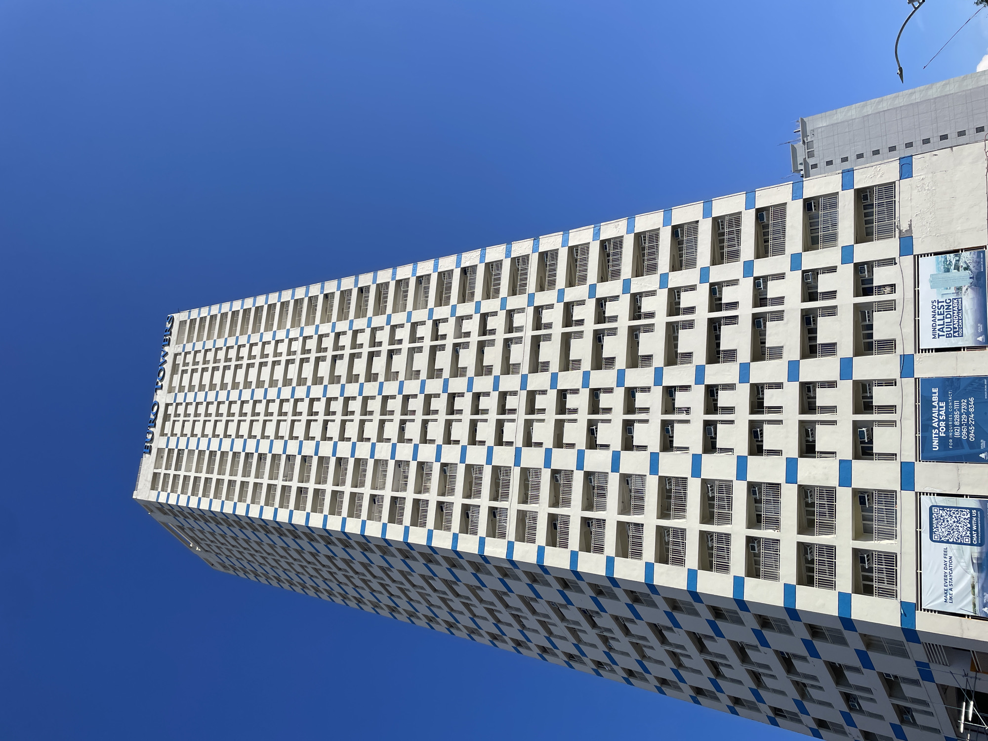 The image shows the Euro Towers building in Davao City, Philippines, against a clear blue sky. The tall, white, residential building features rows of balconies with white railings and blue accents on the facade. The building has "EURO TOWERS" written in blue letters at the top. Various advertisements are visible at the base of the building, including one that reads "UNITS AVAILABLE FOR SALE" with contact numbers. Another advertisement features a QR code and the text "MAKE EVERY DAY FEEL LIKE A STAYCATION. CHAT WITH US." A smaller, lighter-colored building is visible to the right of the Euro Towers, and a streetlamp is also present. The perspective is from a low angle, looking up at the towering structure.